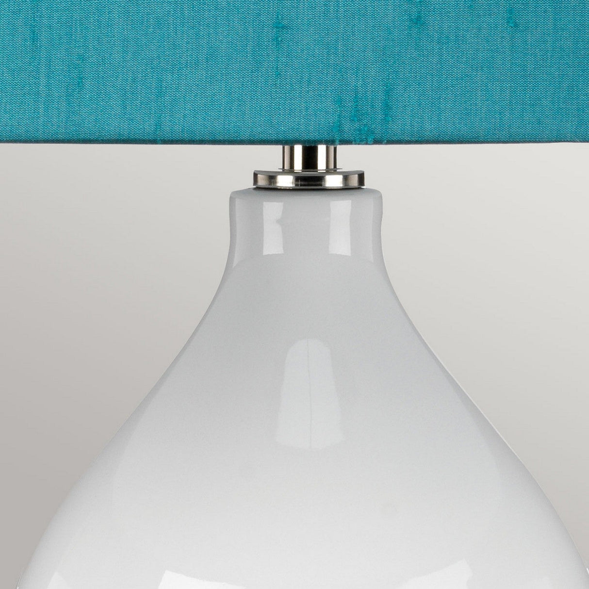 Close-up of the Isla 1 Light Table Light in white ceramic with a glossy finish. It features a teal shade, partially visible at the top, and is connected by a silver metal accent to its ceramic base. The background showcases a plain light gray wall.