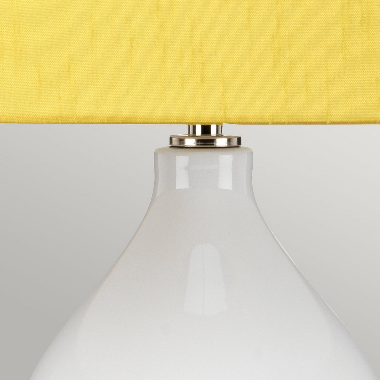 A close-up of the Isla 1 Light Table Light in white and yellow reveals its smooth, glossy ceramic base complemented by a silver-tone neck and a vibrant yellow shade, set against a plain, light gray background.