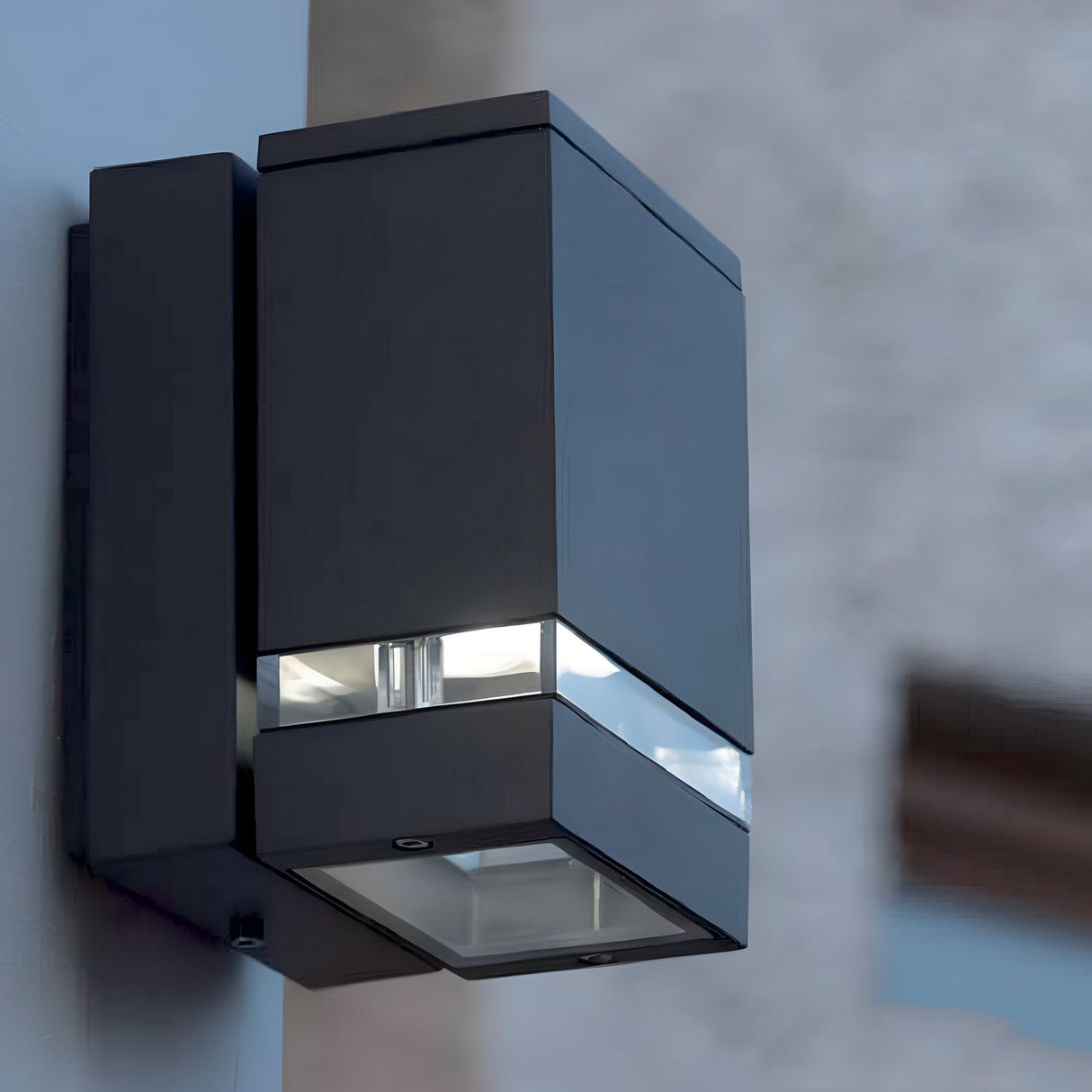 The Jannik 1 Light Outdoor LED Wall Light in graphite, with its modern and minimalist rectangular design, is mounted on a wall. Its small illuminated section indicates that the energy-efficient light is turned on. The blurred background enhances the focus on its sleek design and IP44 weather-resistant rating.