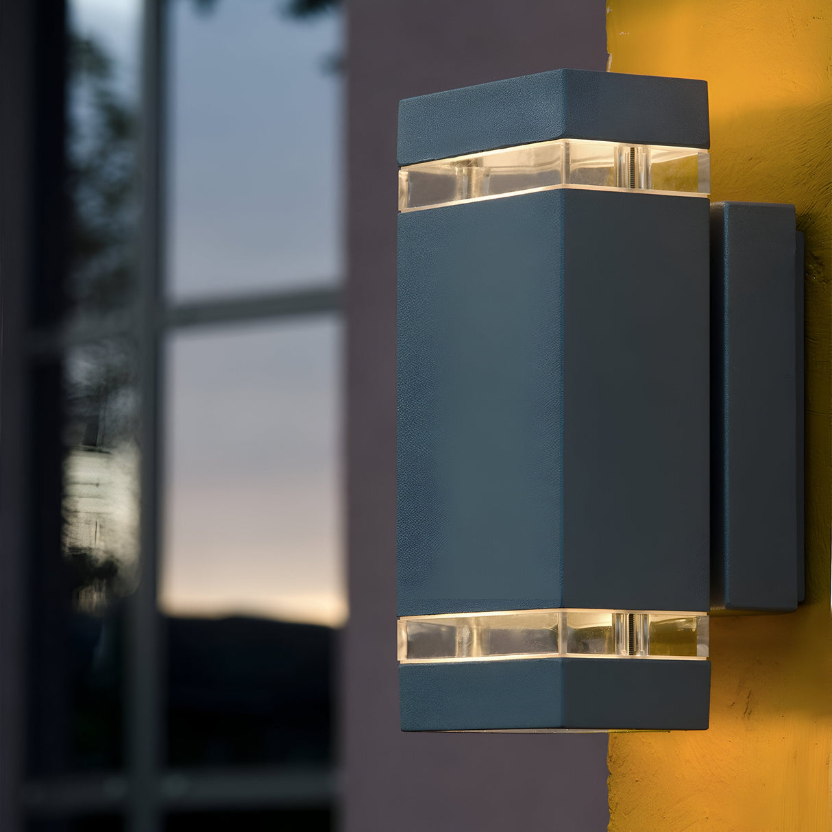 The Jannik 2 Light Outdoor LED Wall Light in Graphite, featuring a sleek geometric design, is mounted on a yellow wall. It's illuminated and casts a warm glow, while a window in the background reflects the dim evening sky, highlighting its energy efficiency and contemporary lighting appeal.