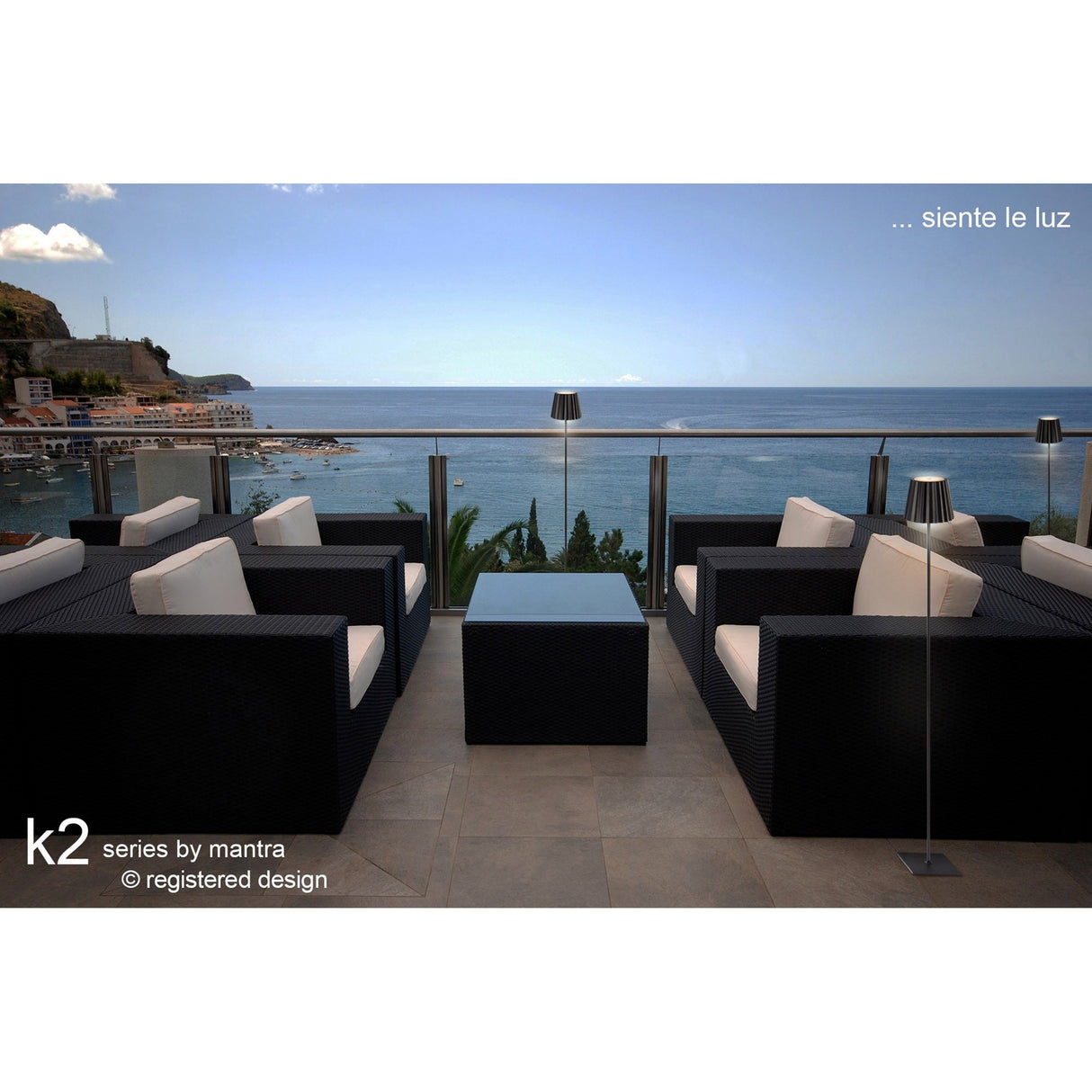 Modern outdoor patio with four black wicker armchairs, a square coffee table, and two K2 LED Battery Outdoor Floor Lamps 2.2W 3000K in Black overlooking the ocean and distant hills under a clear blue sky. Text reads “k2 series by mantra” and “…siente le luz.”.