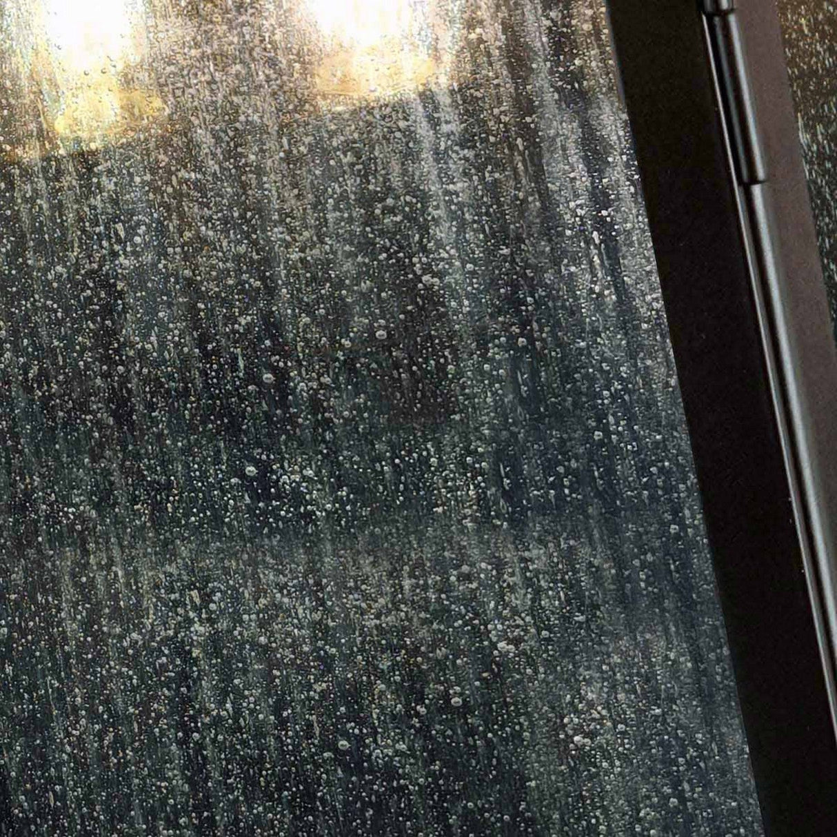 Raindrops slide down a window, forming a textured, watery pattern. The light outside filters through the droplets, casting a warm, amber hue akin to the Kendal 2 Light Outdoor Wall Light in Old Bronze, which stands out against the cool, rainy atmosphere. A slight glimpse of a dark frame appears on the right.