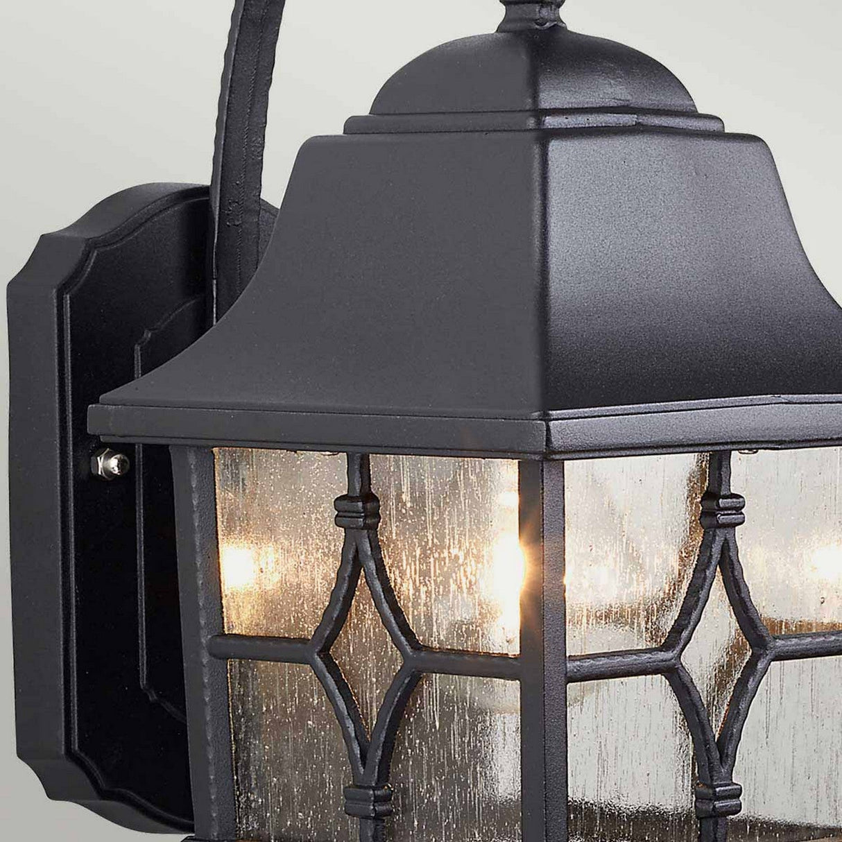 Close-up of the Kent 1 Light Outdoor Wall Light - Black, showcasing its classic design with a sleek black metal finish. The lantern features clear glass panels adorned with decorative geometric patterns and houses a lit bulb that emits a warm glow. Mounted on a wall bracket, it offers both style and durability with an IP44 rating.