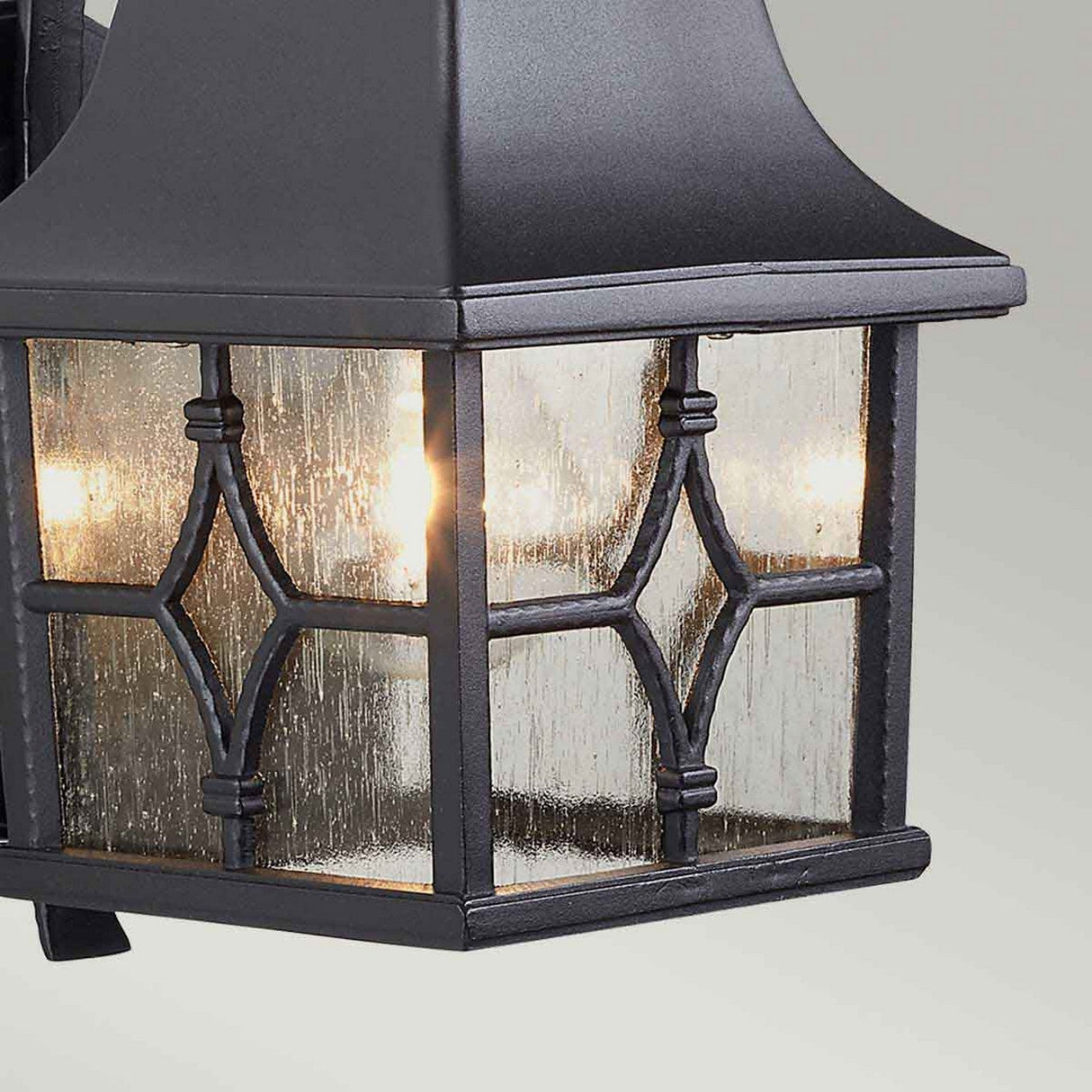 A detailed view of the Kent 1 Light Outdoor Wall Light - Black illustrates its elegant design with transparent glass panels and metal grid patterns, all in a sleek black finish. It emits a warm glow from two visible light bulbs, enclosed in an IP44 rated weather-resistant frame.