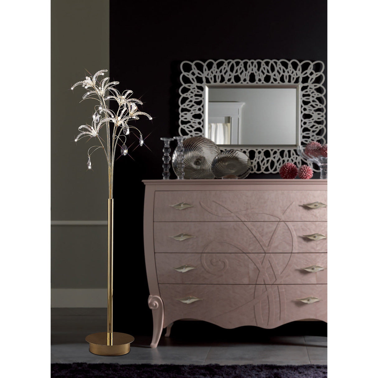 A modern room with the Kenzo 6 Light Crystal Floor Lamp in French Gold, featuring decorative crystal leaves, a light pink ornate dresser topped with sculptural decor, a large mirror, all set against a black wall.