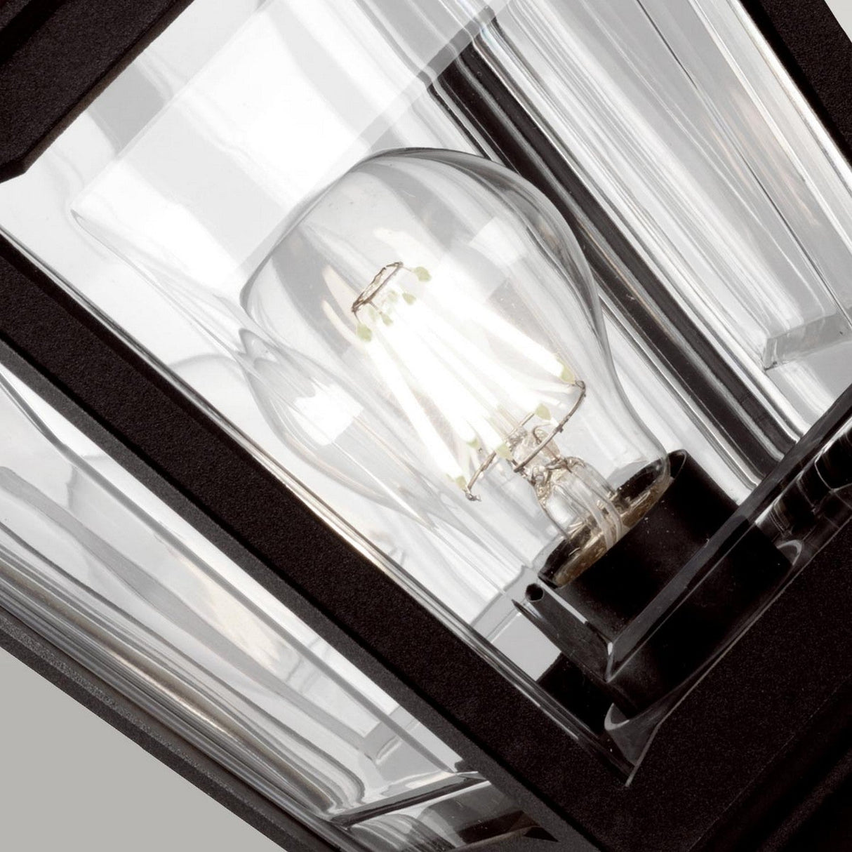 Close-up of the Kerry 1 Light Pedestal/Porch - Black, showcasing a clear bulb enclosed within its black-framed glass panels. The bulb gives off a soft, warm glow, while this corrosion-proof lantern with symmetrical panels and sharp angles adds a modern touch to outdoor spaces against a neutral backdrop.