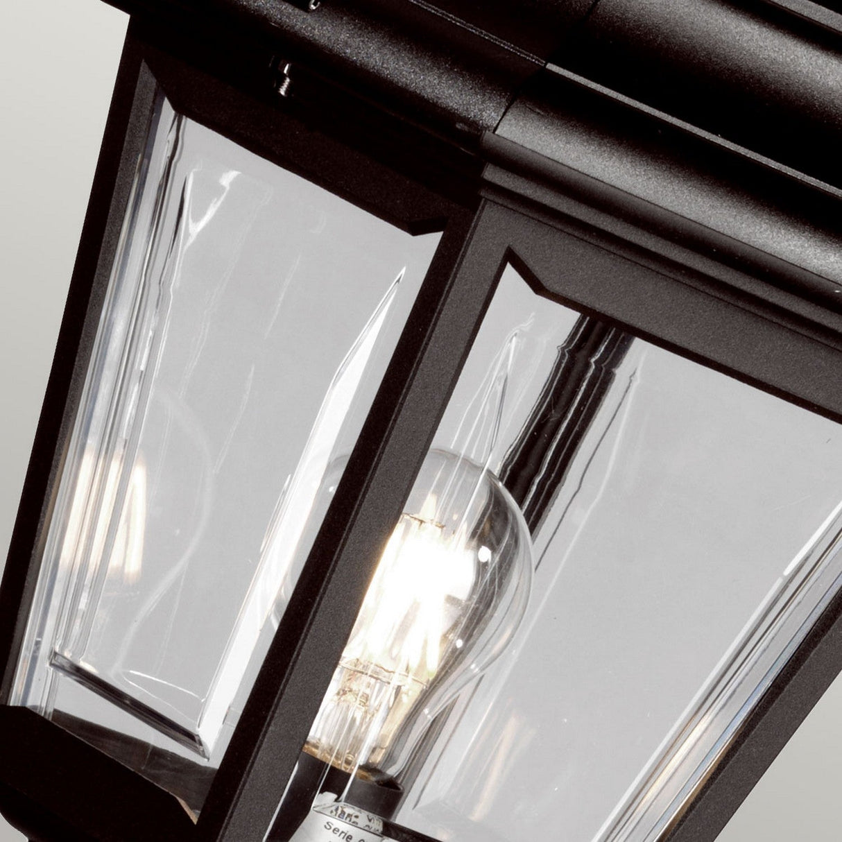 A close-up view of the Kerry 1 Light Pillar Light - Black reveals its modern design with straight lines and a clear glass panel that displays a lit bulb. This outdoor lantern combines sleek style with durable, impact-resistant materials for long-lasting outdoor lighting.