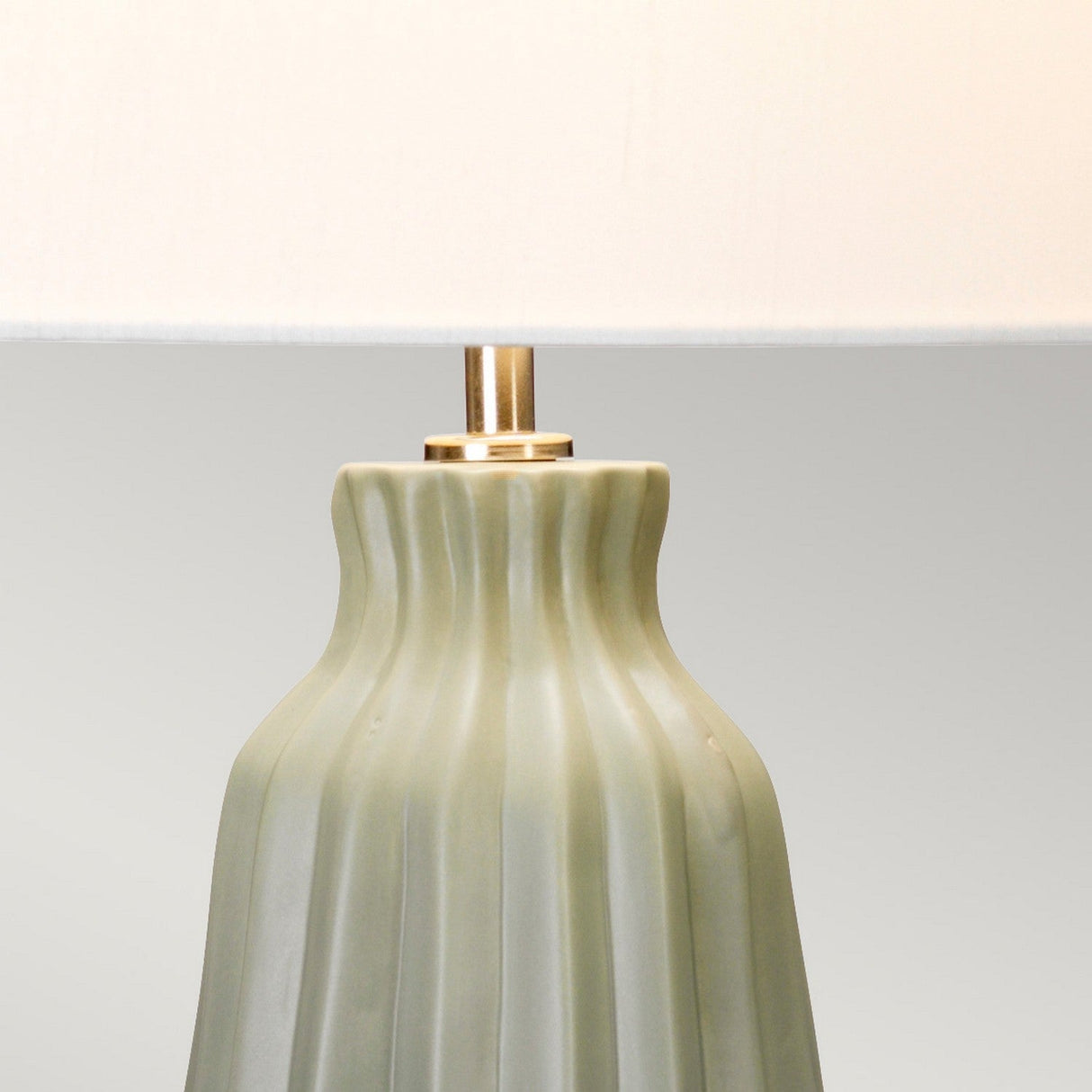 The Kew 1 Light Table Lamp in sage green showcases a botanical-inspired design with a smooth, ridged, cylindrical ceramic base. It is completed with a white lampshade, offering a simple and elegant appearance against the plain light gray background.