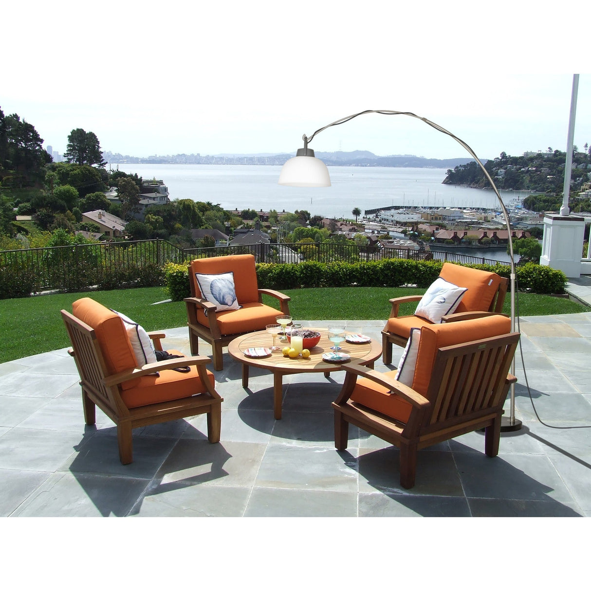 Four wooden patio chairs with orange cushions circle a round table topped with drinks and snacks on a stone patio overlooking water. The black Kinke Floor Lamp With Hook arches overhead, casting a warm glow on the scene.