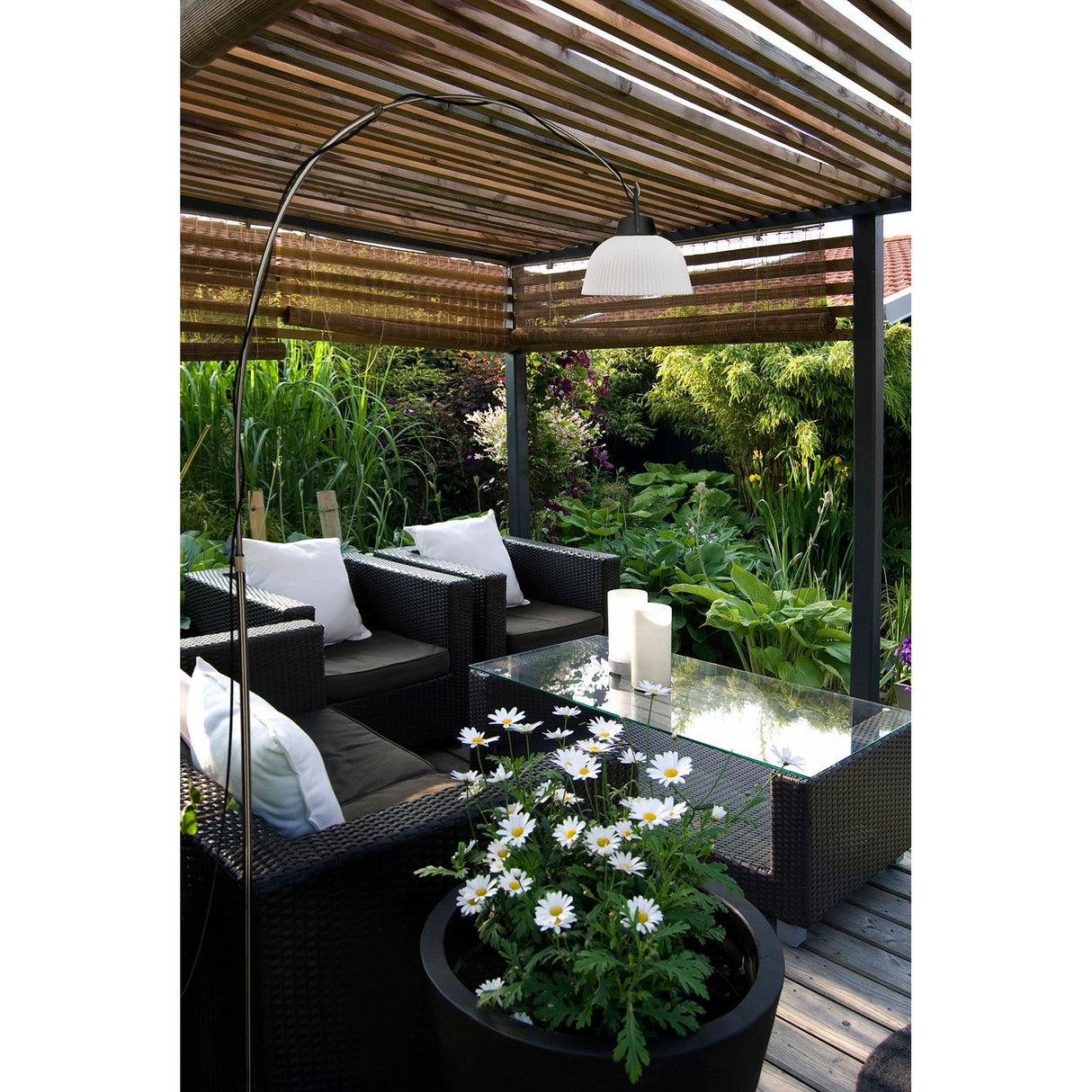 Outdoor patio with black wicker furniture, white cushions, a glass coffee table with candles, a potted daisy plant, and the sleek Kinke Floor Lamp With Hook - Black beside the seating area. Lush greenery surrounds the space under a slatted pergola.