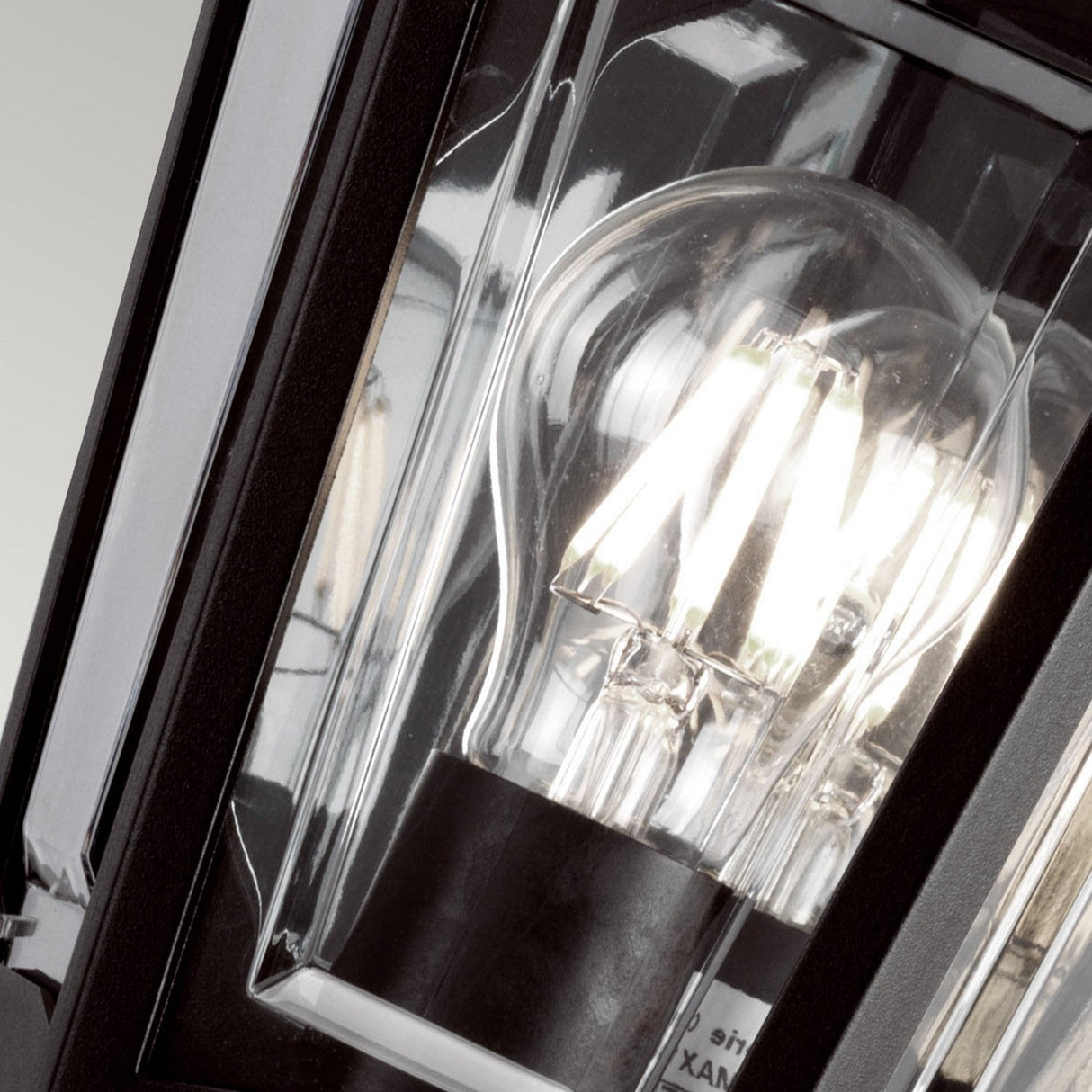 The Kinsale-1 1 Light Outdoor Wall Light - Black features a clear glass light bulb encased in a weather-resistant, black-framed glass enclosure. When lit, the bulb emits a bright glow with reflections visible on the corrosion-proof glass surfaces.