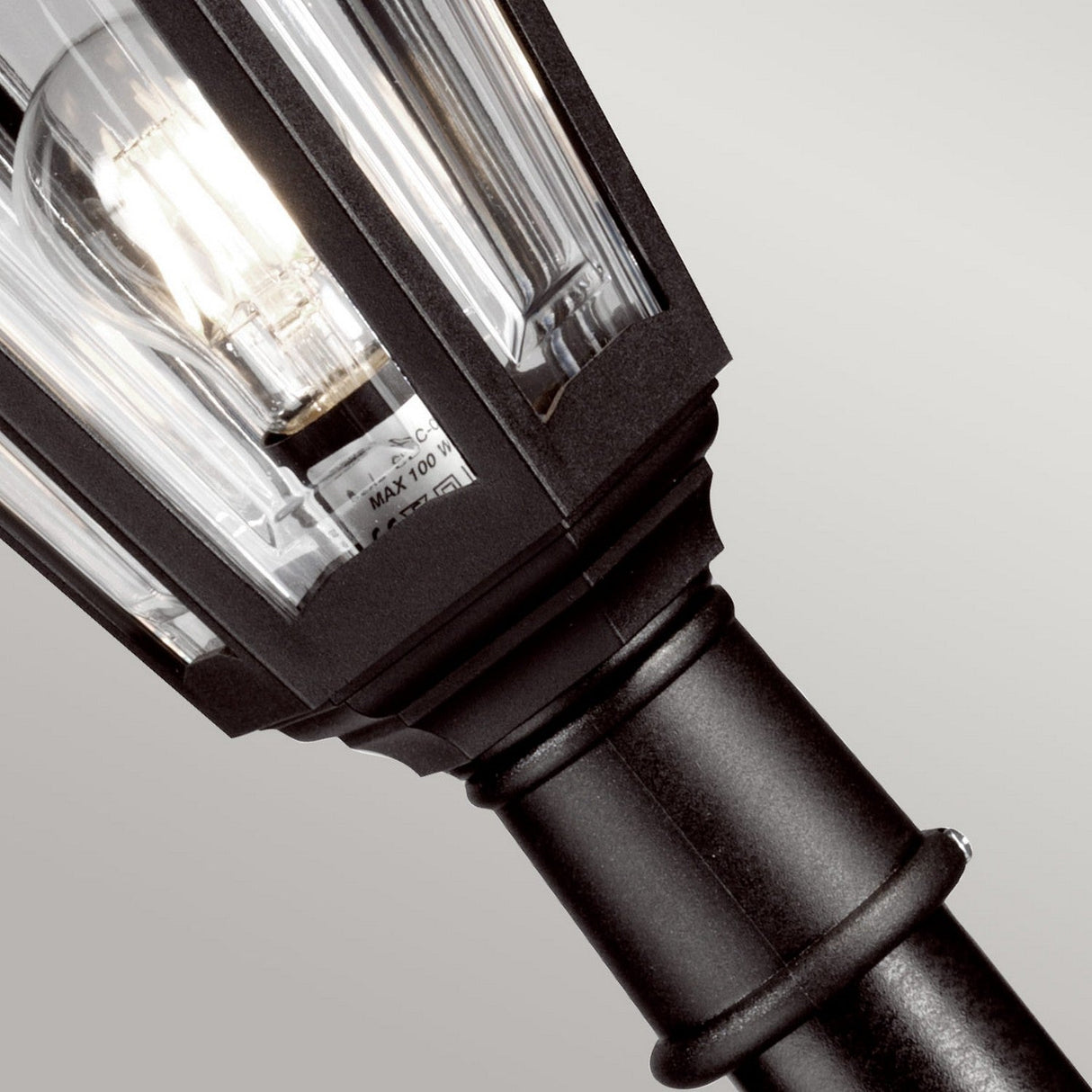 A detailed view of the Kinsale 1 Light Pillar Light in black, highlighting a clear glass enclosure that reveals a visible light bulb. This timeless outdoor lighting fixture, designed with angular edges and corrosion resistance, is displayed against a simple grey background.