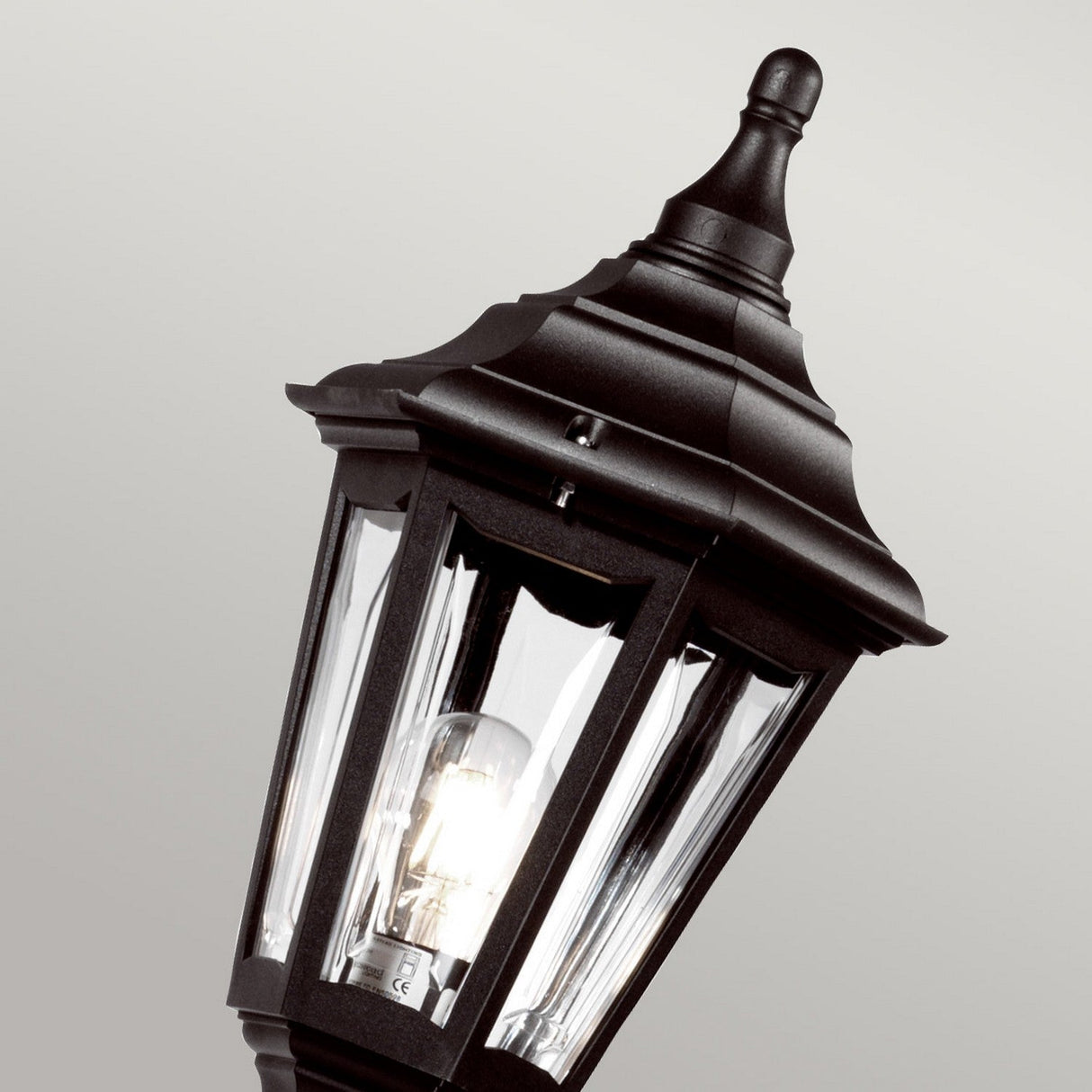 The Kinsale 1 Light Lamp Post in Black features a corrosion-proof design and a vintage aesthetic with its clear glass casing and illuminated bulb, making it an ideal choice for coastal lighting. Set against a simple, light gray background, it provides dependable outdoor illumination with a touch of timeless elegance.