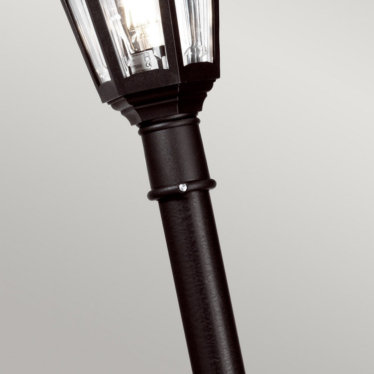 The Kinsale 1 Light Lamp Post in black presents a vintage-style appearance with a hexagonal lantern top, providing corrosion-proof outdoor lighting against a plain grey background. Featuring a textured surface and an exposed bulb inside the lantern, it's ideal for coastal lighting needs.