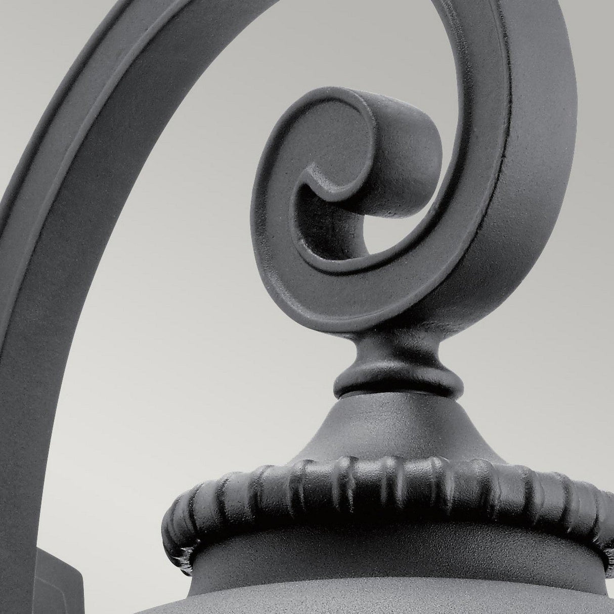 Close-up of the Admirals Cove Outdoor 1 Light Large Wall Lantern in black, showcasing its ornate metal scroll design inspired by Tudor aesthetics. It features a curved loop with a textured edge against a plain, light gray backdrop, making it perfect for outdoor lighting.