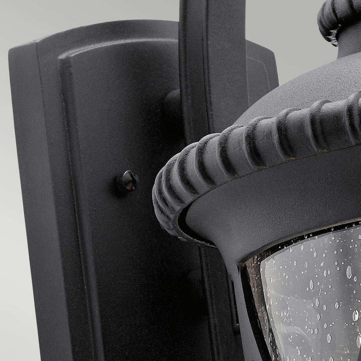 A close-up of the Admirals Cove Outdoor 1 Light Large Wall Lantern in black, mounted on a wall. The lantern showcases a Tudor-inspired design with glass panels speckled with water droplets and features a textured, rope-like detail around its top. It is anchored by a large rectangular wall mount with a smooth finish.
