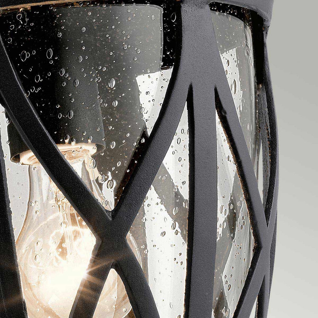 Close-up of the Admirals Cove Outdoor 1 Light Large Wall Lantern - Black, featuring a Tudor-inspired design with a clear, rain-speckled glass cover and a black metal lattice. Inside, a glowing bulb casts warmth, creating a striking contrast against the water droplets on this elegant lantern.