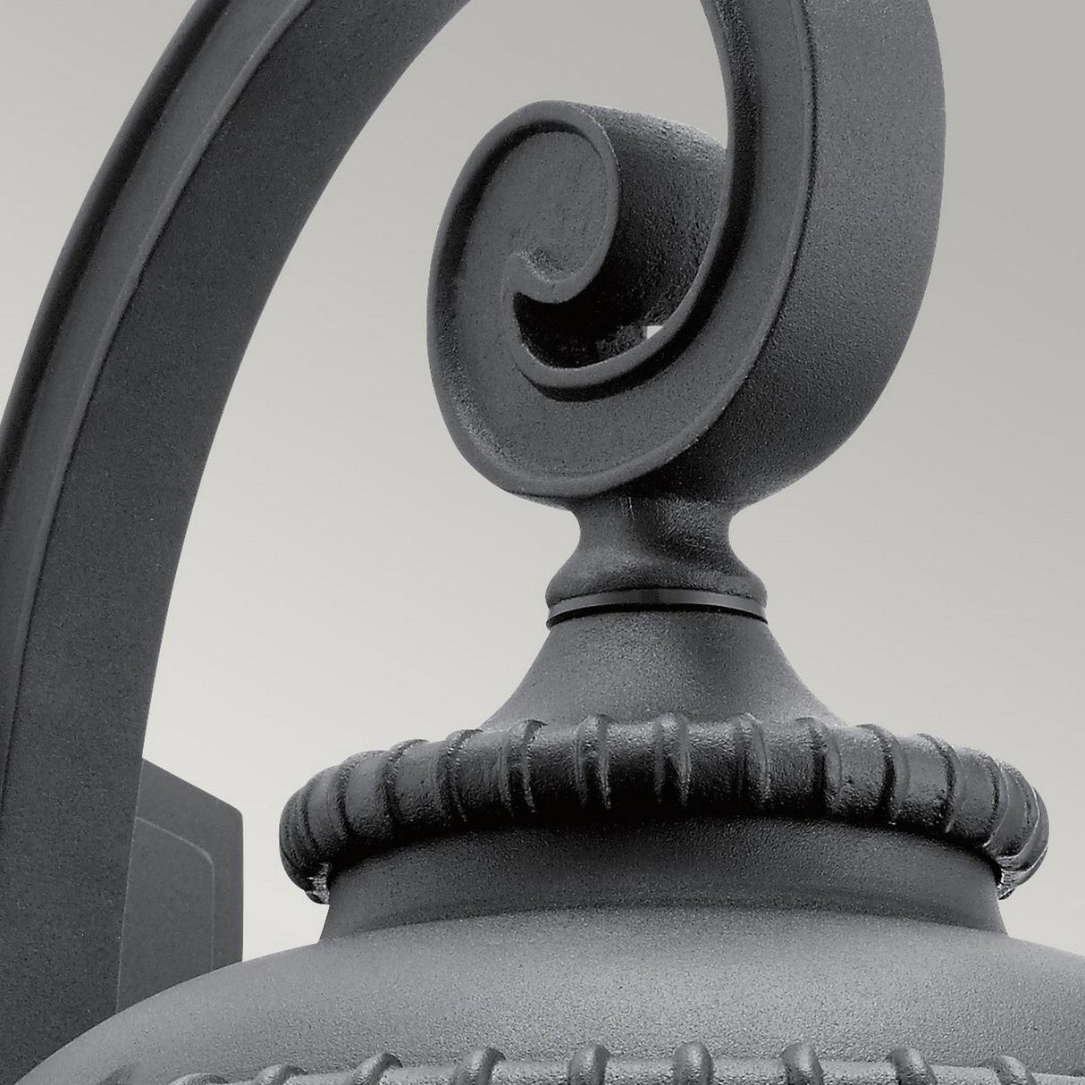 A close-up of the Admirals Cove Outdoor 1 Light Medium Wall Lantern showcases its intricate scrollwork and ribbed design in vintage style. This Tudor-inspired fixture, featuring a textured black finish, stands out against a plain background.