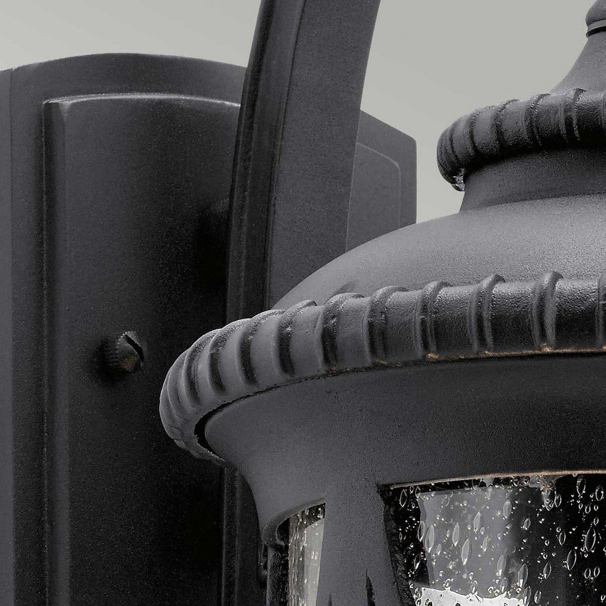 Close-up of the Admirals Cove Outdoor 1 Light Medium Wall Lantern - Black, attached to a wall. The Tudor-inspired lantern features a textured glass pane with water droplets that hint at exposure to rain and boasts detailed metalwork with a curved top design.