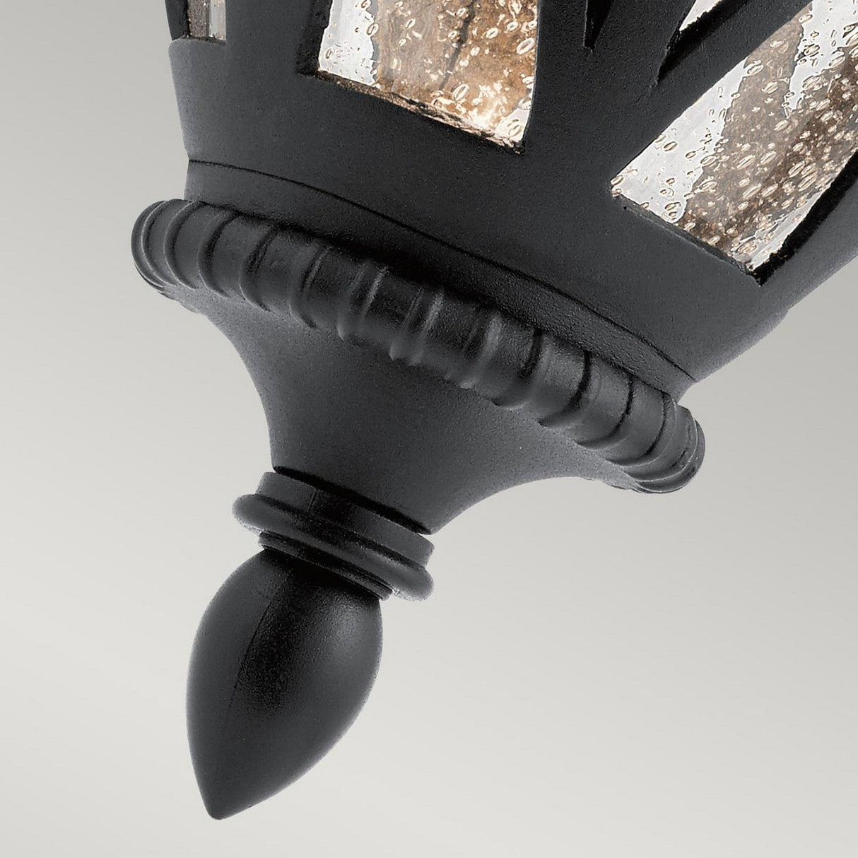 Close-up of the Admirals Cove Outdoor 1 Light Medium Wall Lantern in black, featuring ornate, Tudor-inspired metalwork. The design is completed with clear, textured glass panels and a pointed finial, set against a plain gray background.