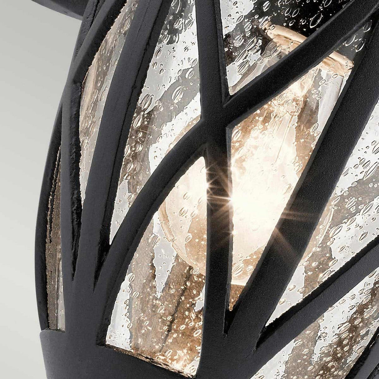 Close-up of the Admirals Cove Outdoor 1 Light Medium Wall Lantern in black, showcasing a Tudor-inspired decorative light fixture. It features an intricate textured black metal lattice design. Behind the lattice, a clear glass shade with a waterdrop texture softly diffuses the light, creating a warm glow reminiscent of an outdoor wall lantern.