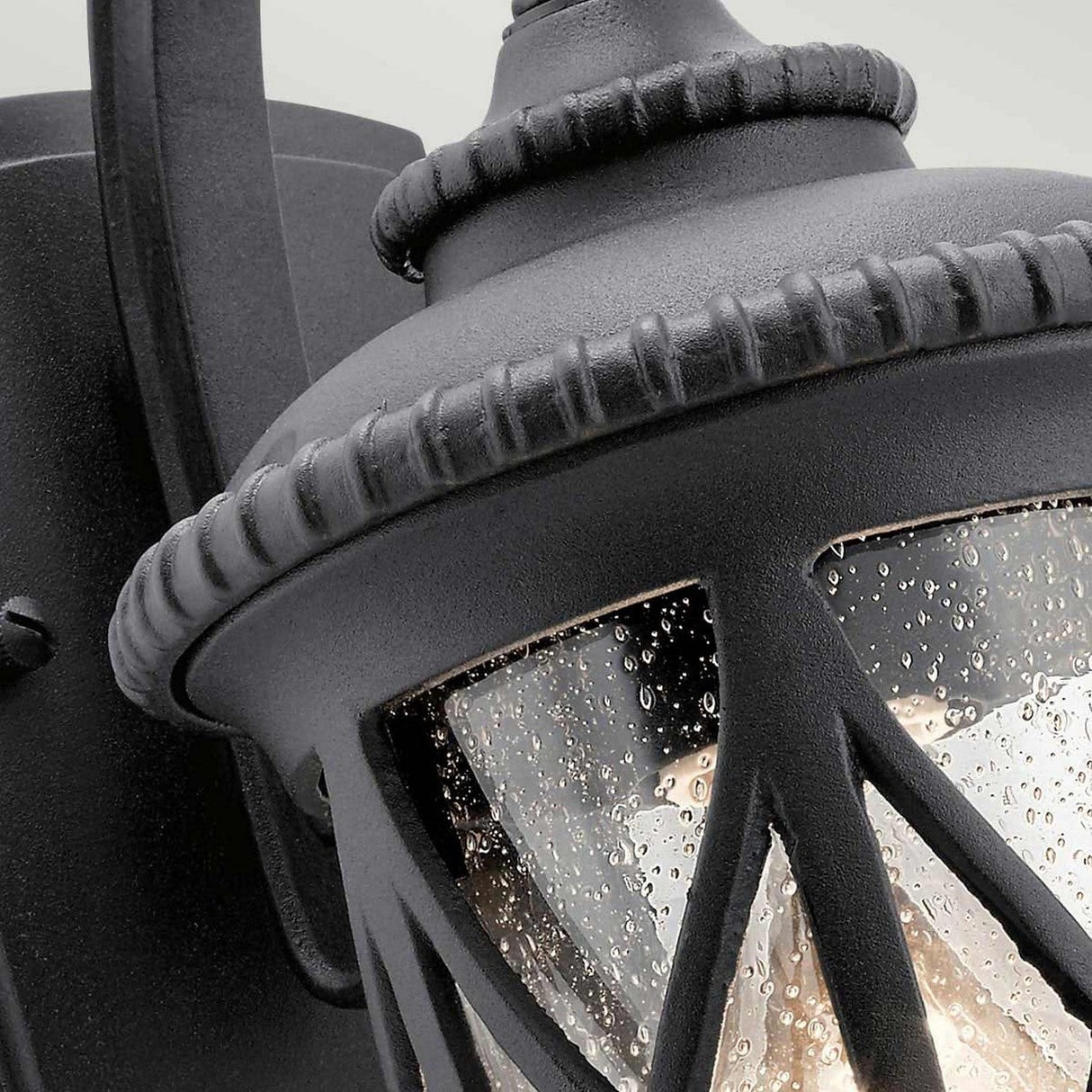 Close-up of the textured Admirals Cove Outdoor 1 Light Small Wall Lantern in black, featuring raindrop-textured glass and intricate details with a curved mounting bracket. The IP44 rated lantern is illuminated, highlighting its classic design.