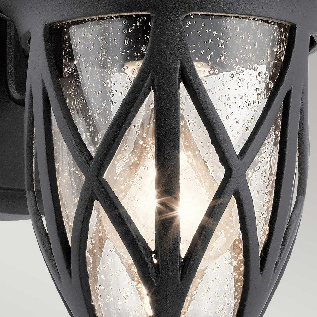 Close-up of the Admirals Cove Outdoor 1 Light Small Wall Lantern in black, featuring a decorative crisscross pattern. The glass is speckled with droplets, while a warm light emanates from the filament bulb inside, providing a soft glow. Designed to be IP44 rated, it elegantly withstands various weather conditions.