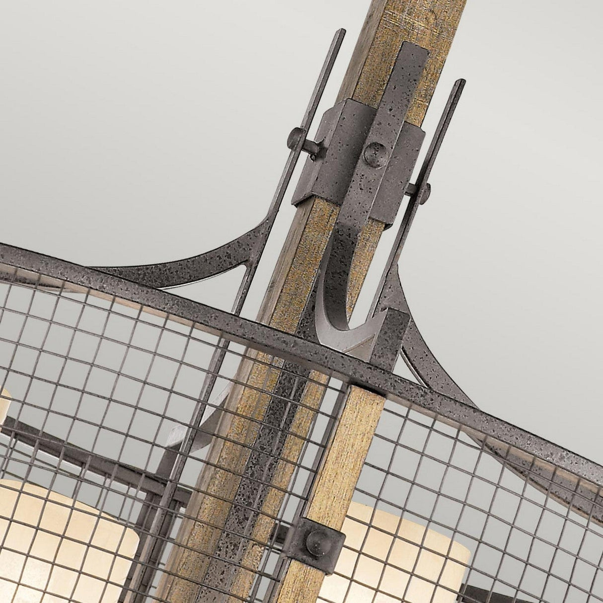 A close-up of the Ahrendale 4 Light Chandelier in Grey showcases its metal and wood structure, featuring a grid-like pattern with ornate metal brackets that exude rustic charm. The design is reminiscent of an anvil iron mesh, incorporating both curved and straight elements centered around a central post, set against a plain light background.