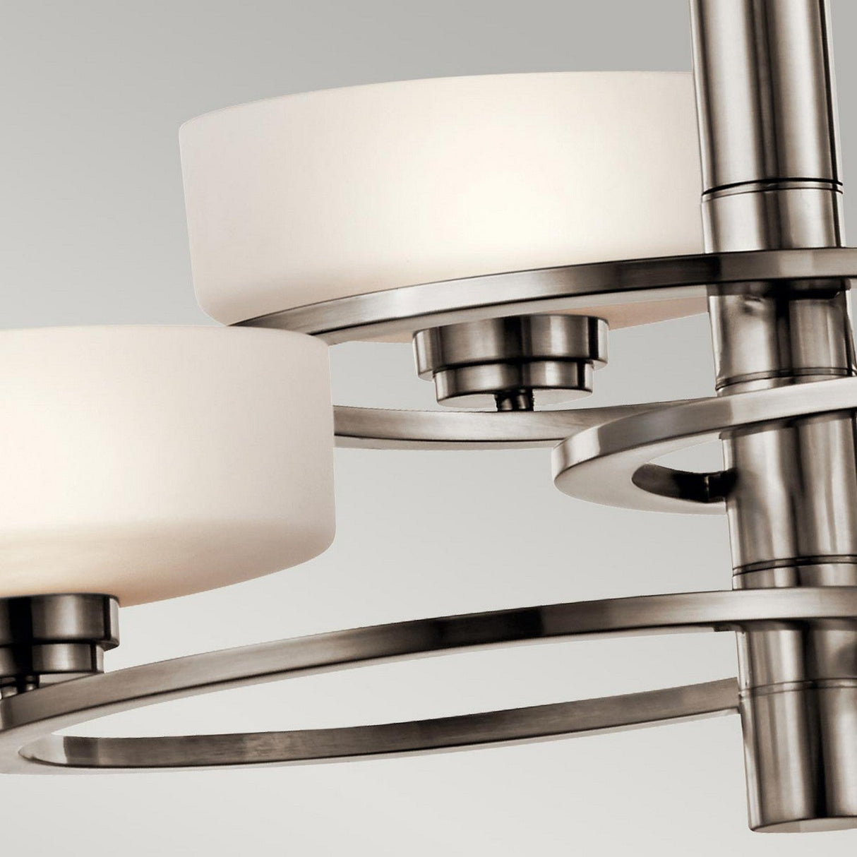 A close-up of the Aleeka 3 Light Chandelier - Silver highlights two frosted glass lampshades and metallic supports against a gray backdrop. This piece exemplifies contemporary lighting with its minimalist design, clean lines, and brushed metal finish—perfect for modern interiors.