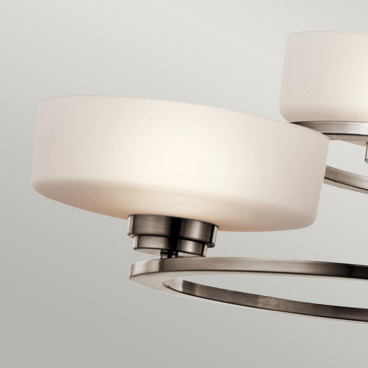 A close-up view of the Aleeka 3 Light Chandelier - Silver, showcasing a modern ceiling light fixture with a brushed metal finish. Ideal for contemporary interiors, it features a circular metal arm and two frosted white glass shades, set against a neutral background.
