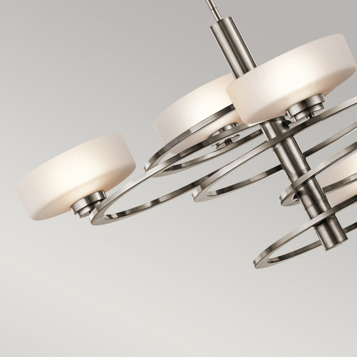 The Aleeka 5 Light Chandelier - Silver hangs against a light gray background, showcasing its modern celestial-inspired design. This elegant fixture features sleek metal rings and round white bulb covers, with multiple circular lights arranged in a spiral formation for a contemporary look.
