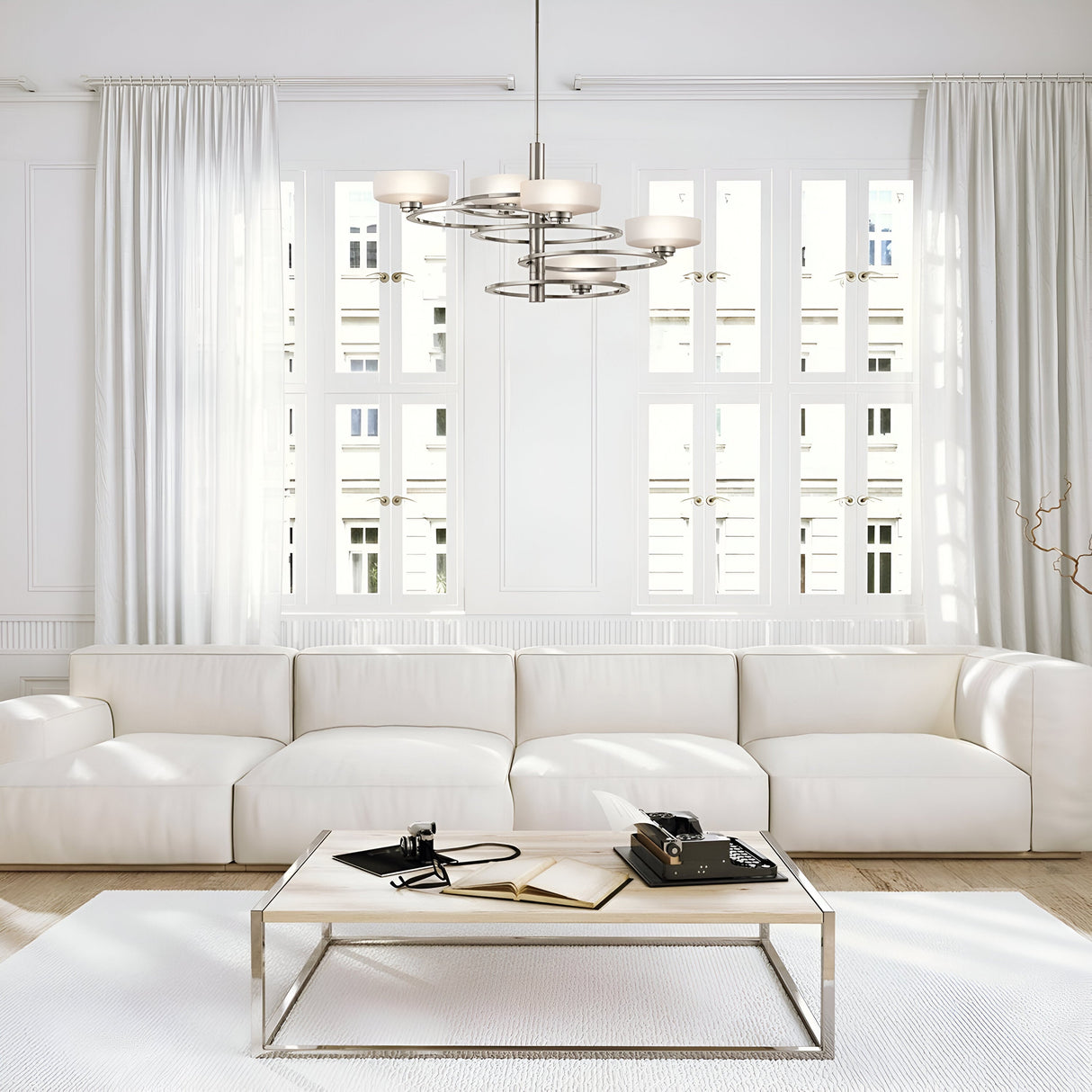 In a bright, minimalist living room that captures celestial inspiration, a large white sectional sofa is complemented by the Aleeka 5 Light Chandelier in Silver and a sleek coffee table. On the table rest a book, phone, and camera. Tall windows with sheer curtains flood the space with natural light, enhancing the contemporary interior design.