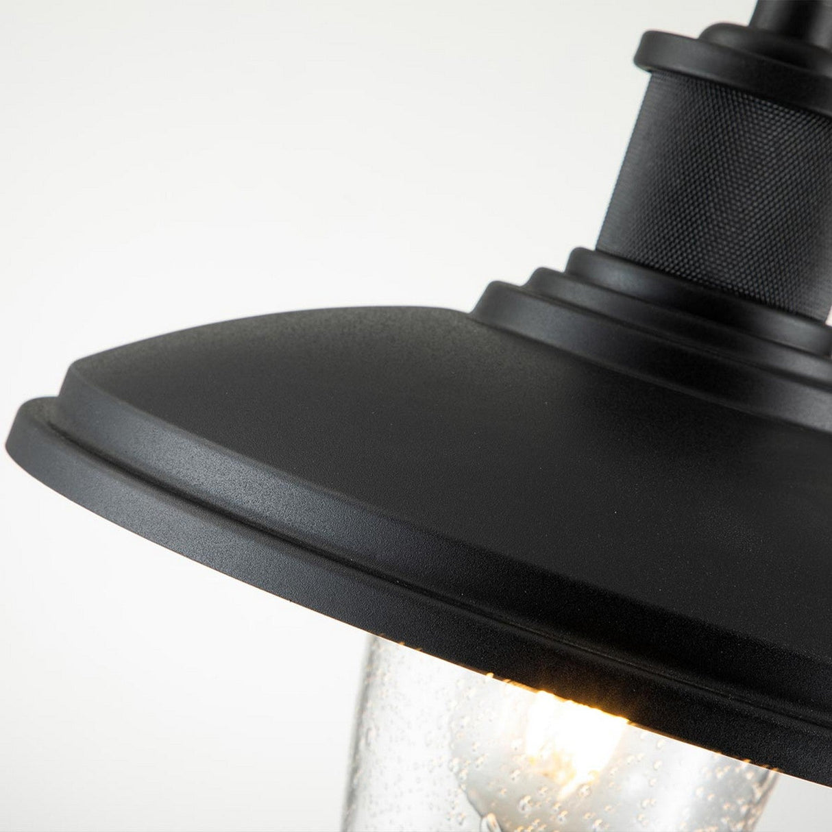 Close-up of the Allenbury Outdoor 1 Light Medium Wall Lantern in black, featuring a weather-resistant design and a dome-shaped top. The glowing IP44-rated light inside emits a gentle glow, and the glass cover beneath showcases a speckled watery texture. The softly lit background highlights the lantern's intricate details.