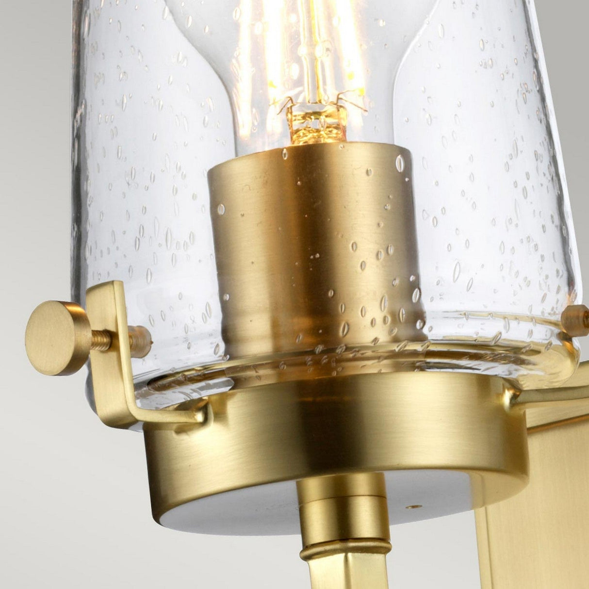 Close-up of the Alton 1 Light Wall Light in brushed brass, featuring a clear glass shade with bubble-like textures. This fixture emits a soft, warm glow that adds an inviting atmosphere and industrial-era charm to your bathroom decor, elegantly complementing a neutral backdrop.