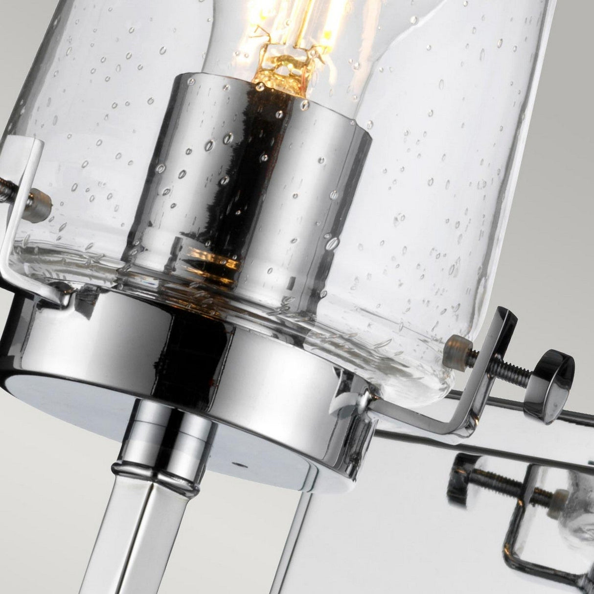 The close-up of the Alton 1 Light Wall Light - Chrome reveals a contemporary fixture featuring a chrome base and a clear, textured glass shade. The exposed bulb casts a warm glow, while the elegant lines and metallic details contribute to its minimalist design, making it an ideal choice for industrial bathroom lighting.