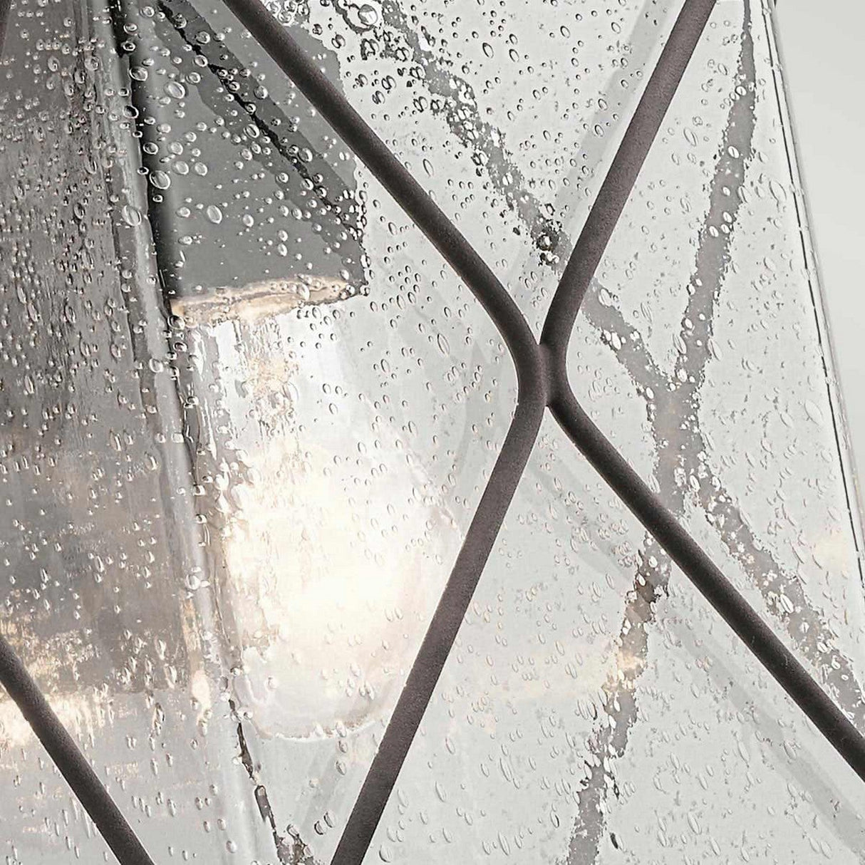 The Argyle Outdoor 1 Light Medium Wall Light in Weathered Zinc showcases a bulb glowing behind seeded glass, highlighted by intersecting dark metal lines. The rain-kissed surface, paired with the weathered zinc finish, offers a textured and geometric appeal.