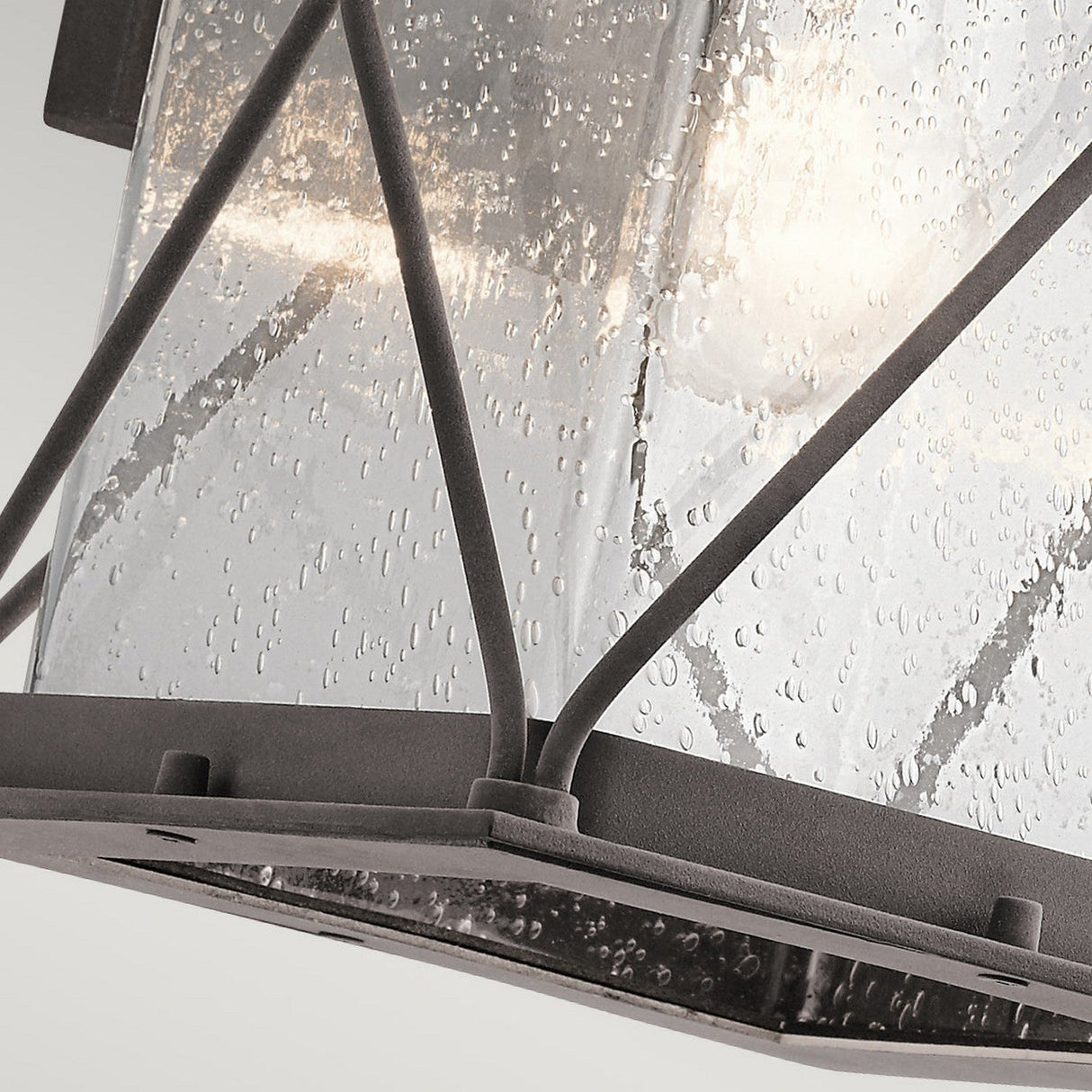 Close-up of the Argyle Outdoor 1 Light Medium Wall Light, featuring clear seeded glass encased in a sleek black metal frame with intersecting bars that emulate a weathered zinc appearance. The interior bulb emits a gentle glow, providing a warm ambiance.