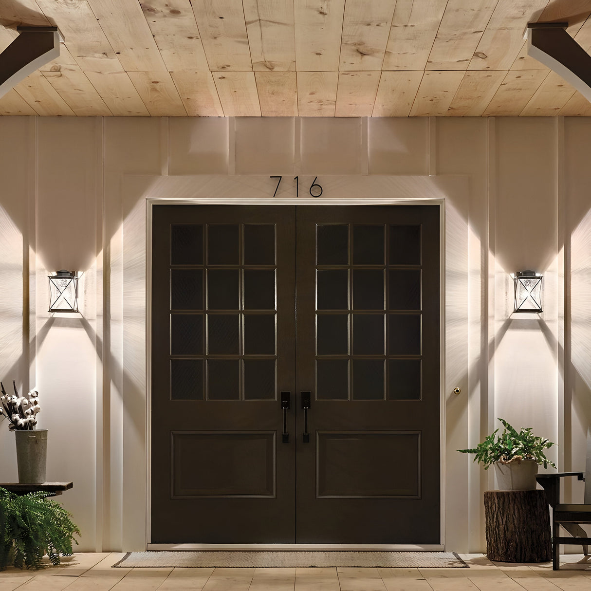 Double front doors featuring seeded glass panes and the number 716 above are complemented by two Argyle Outdoor 1 Light Medium Wall Lights in weathered zinc. The ceiling showcases a wooden finish, while a small potted plant with a fern and a tree stump adorn each side.