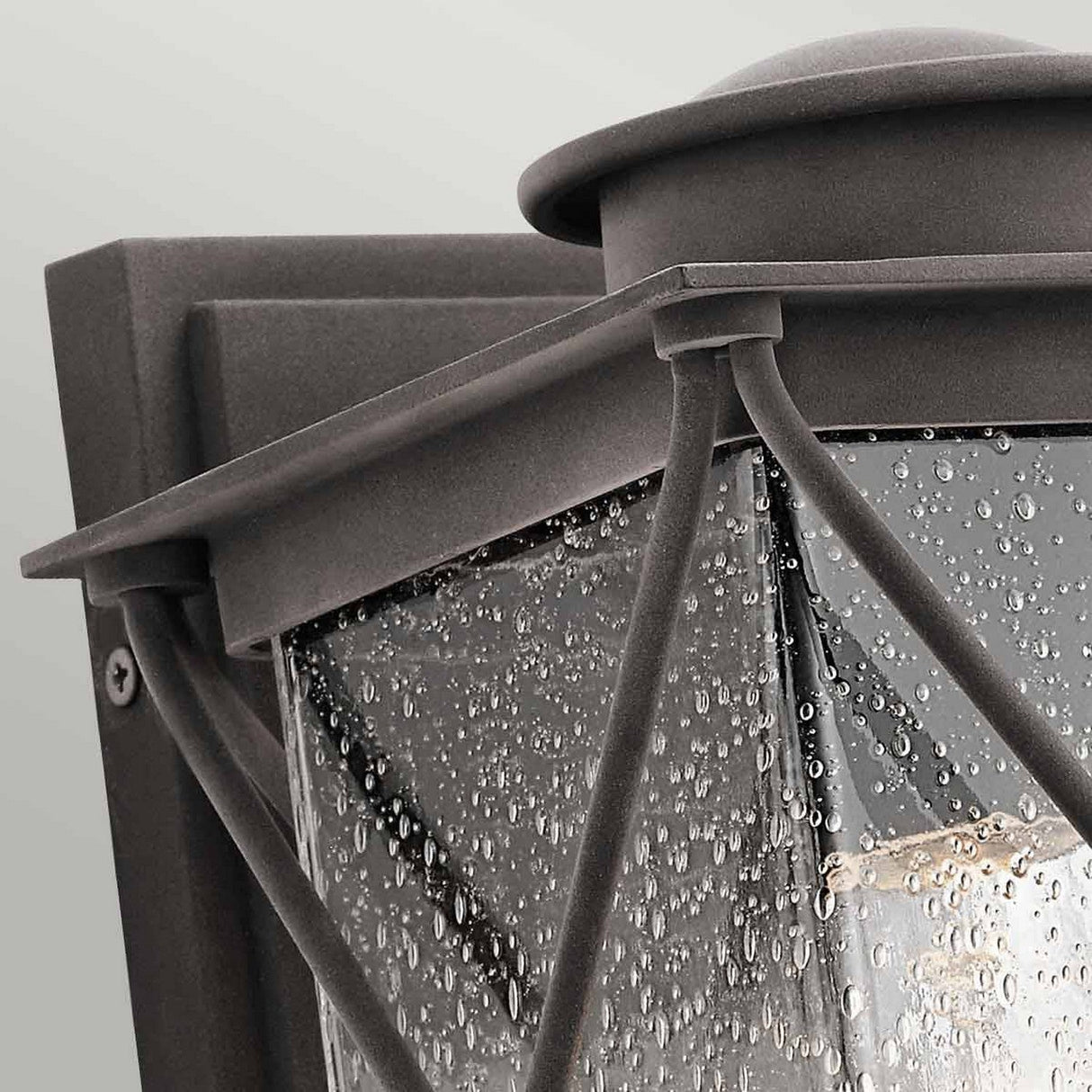 Close-up of the Argyle Outdoor 1 Light Small Wall Light - Weathered Zinc, mounted on a wall. This black outdoor lantern features seeded glass panels with raindrop details and an internal light bulb, all enhanced by decorative crisscross metal bars.