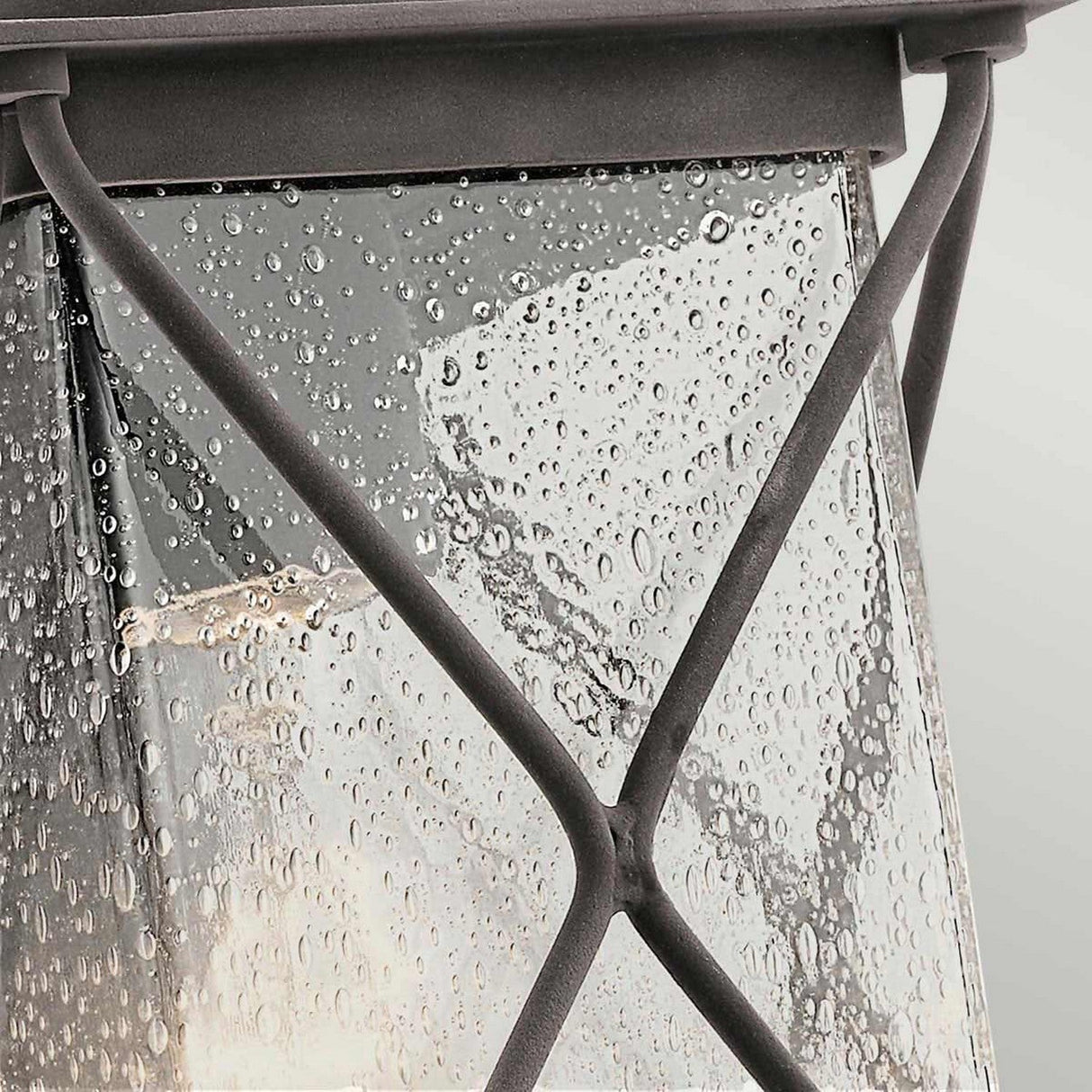 Close-up of an outdoor wall light named "Argyle Outdoor 1 Light Small Wall Light - Weathered Zinc," featuring a black metal lantern with raindrop-textured, seeded glass panels. The internal light emits a warm glow behind the crisscross metal design on the glass surface.