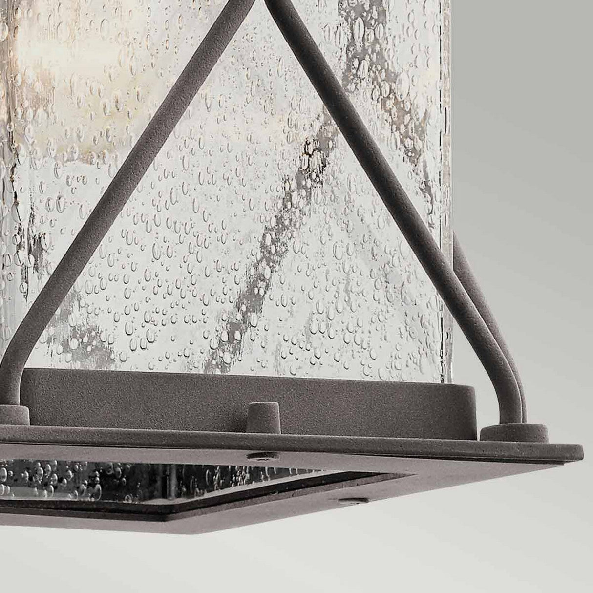 Close-up of the Argyle Outdoor 1 Light Small Wall Light in weathered zinc, showcasing its modern design with a textured seeded glass shade featuring droplets on the surface. The light is complemented by diagonal support bars and a flat square base, all set against a plain background.