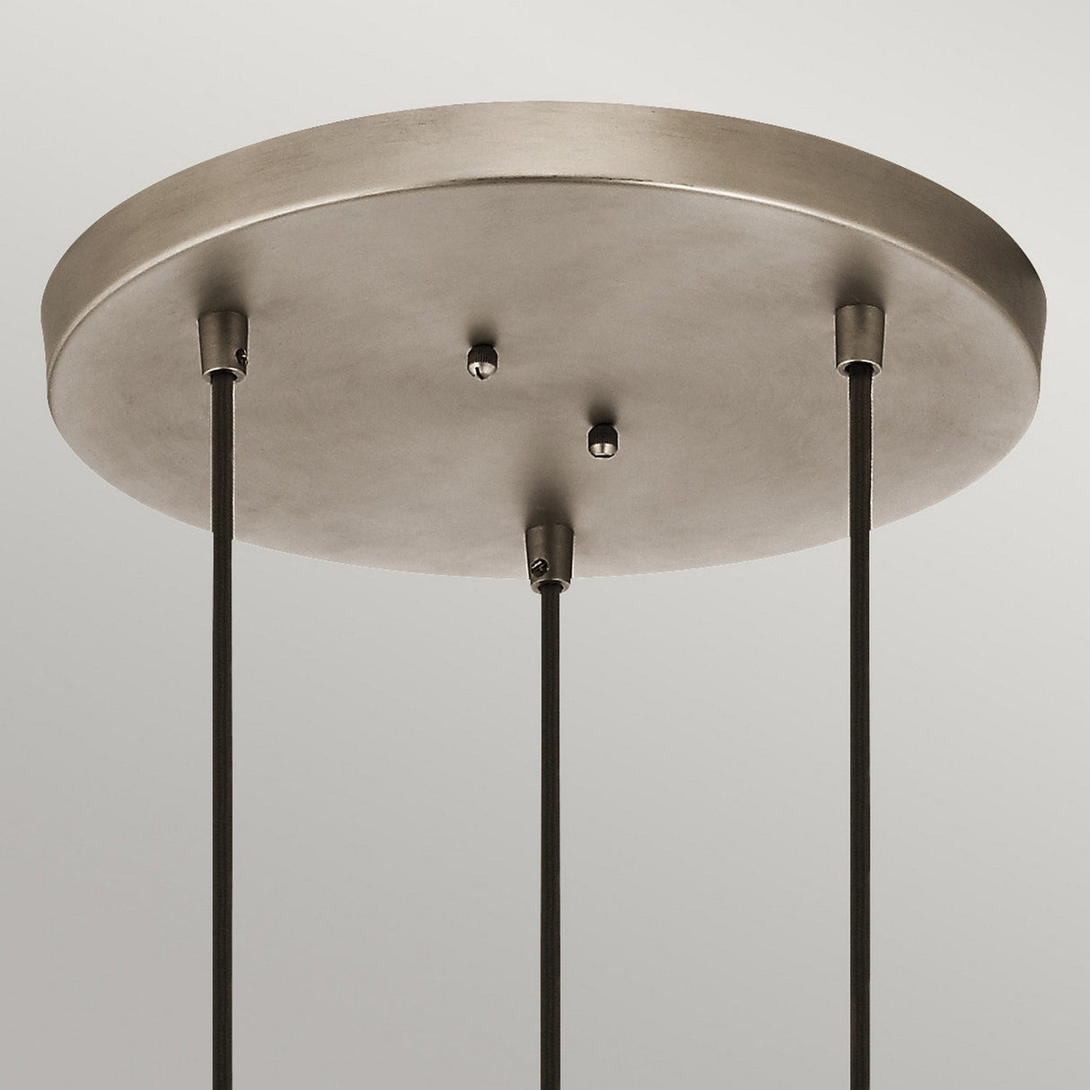 The Asher 3 Light Pendant - Silver showcases an elegant circular design with a round metallic chandelier base and three black cords. Its industrial-inspired aesthetic is highlighted by a brushed silver finish, beautifully mounted on a light gray ceiling.