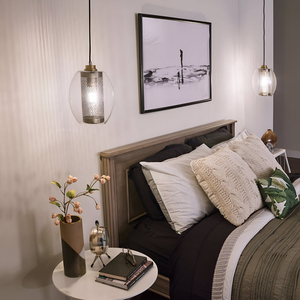 A cozy bedroom highlights modern design with a neatly made bed adorned with green and beige pillows and blankets. Two Asher 1 Light Mini Pendants in silver illuminate the space. A round side table with a plant and books is placed beside the bed, while monochrome artwork decorates the wall above the headboard.