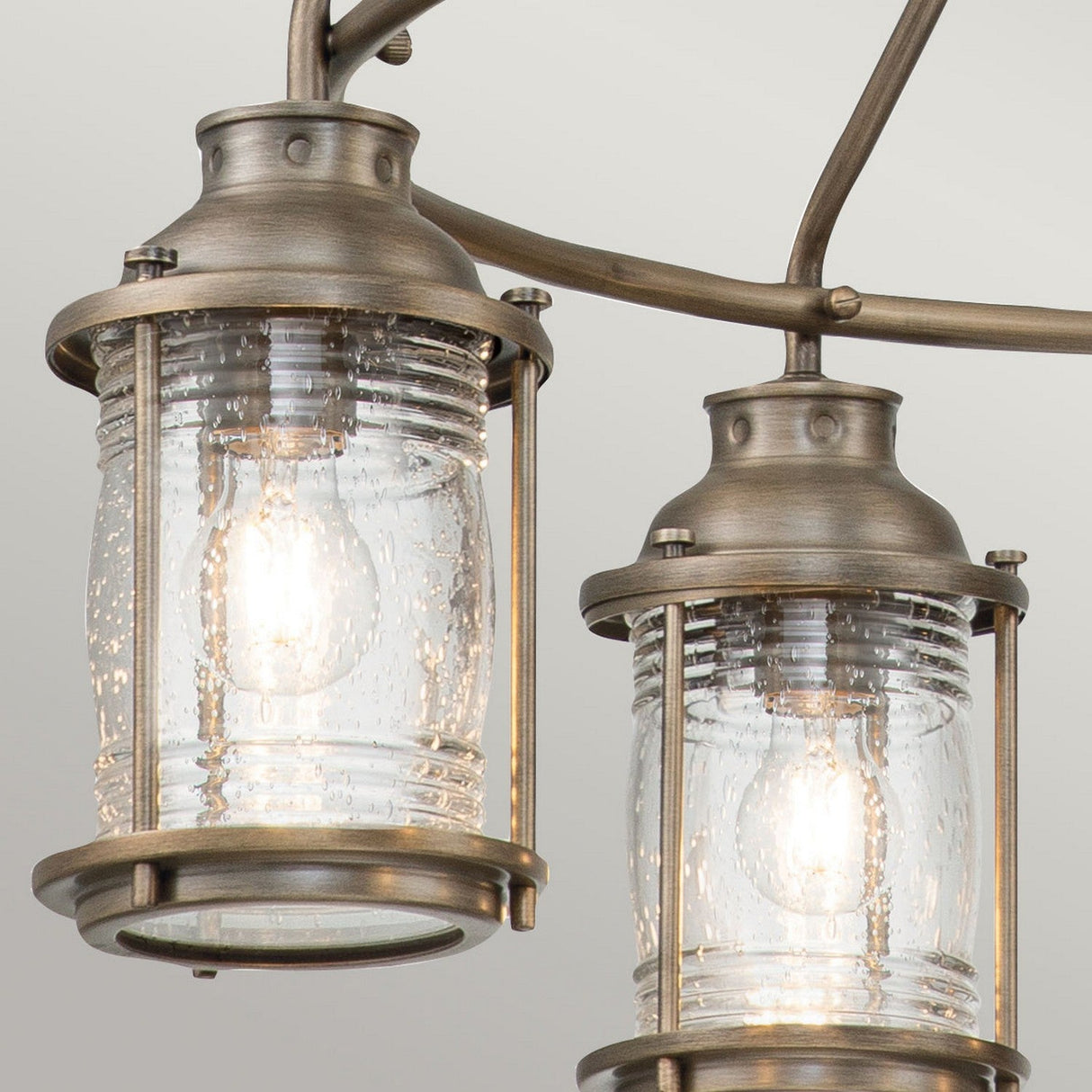 Close-up of two rustic-style lights from the Ashland Bay Outdoor 5 Light Chandelier, featuring clear, seeded glass shades and burnished bronze frames with visible bulbs suspended from a curved metal fixture.