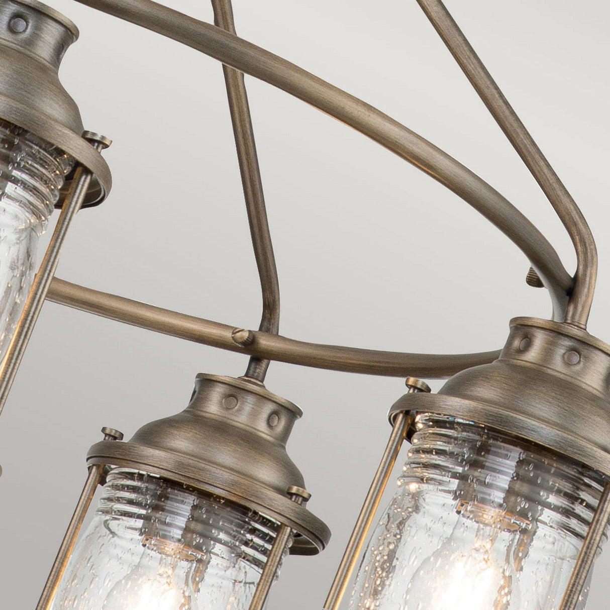 Close-up of the Ashland Bay Outdoor 5 Light Chandelier, showcasing five cylindrical seeded glass light fixtures. The fixtures feature a burnished bronze finish and contain visible filament bulbs, gracefully suspended from curved metal arms against a neutral background.