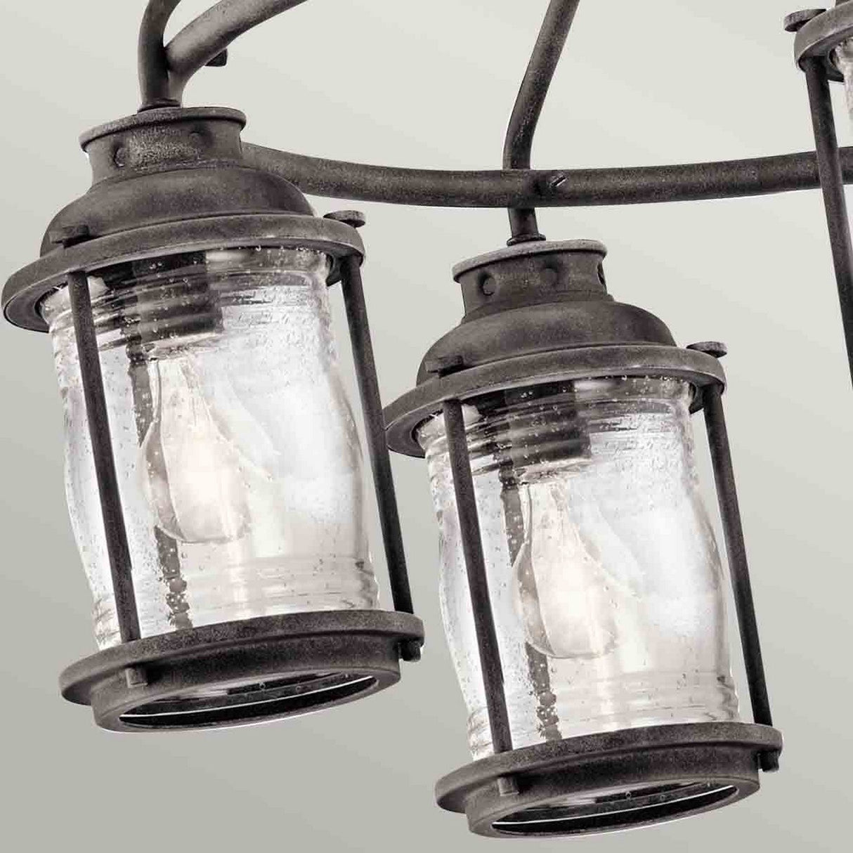 Close-up of the Ashland Bay Outdoor 5 Light Chandelier in weathered zinc, showcasing its two vintage-style metal and glass lanterns with clear light bulbs. The plain, light gray background enhances its outdoor chandelier aesthetic.