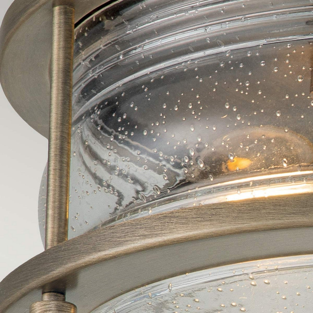 Close-up of the Ashland Bay Outdoor 2 Light Ceiling Flush, featuring a burnished bronze finish. The glass, textured with small air bubbles, encloses an Edison-style bulb that emits a warm glow, while vertical metal rods enhance its industrial design.