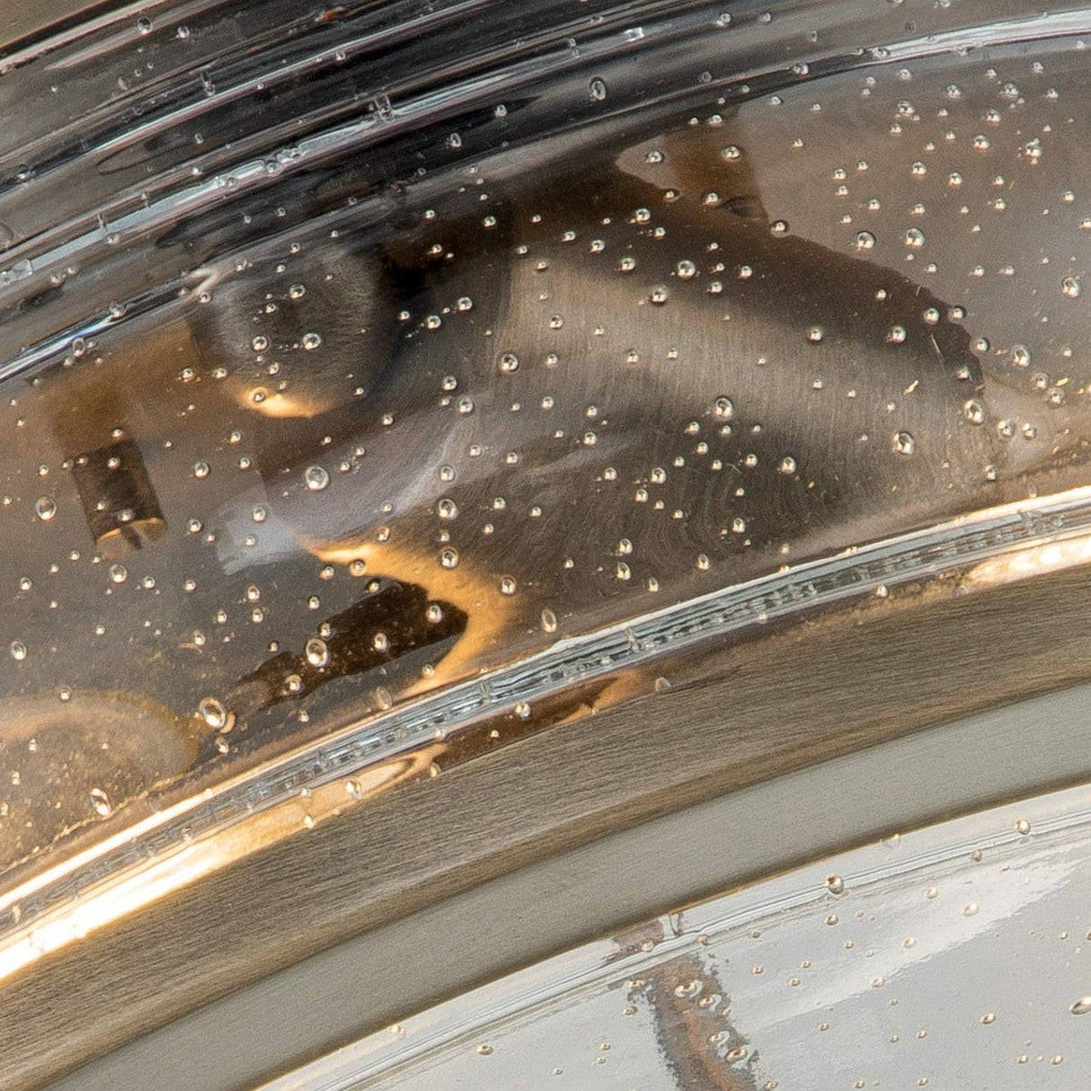 A close-up of the Ashland Bay Outdoor 2 Light Ceiling Flush in Burnished Bronze, featuring water droplets on its sleek surface. The image highlights the curved lines and lustrous texture of the metal, with light reflecting off the scattered droplets.