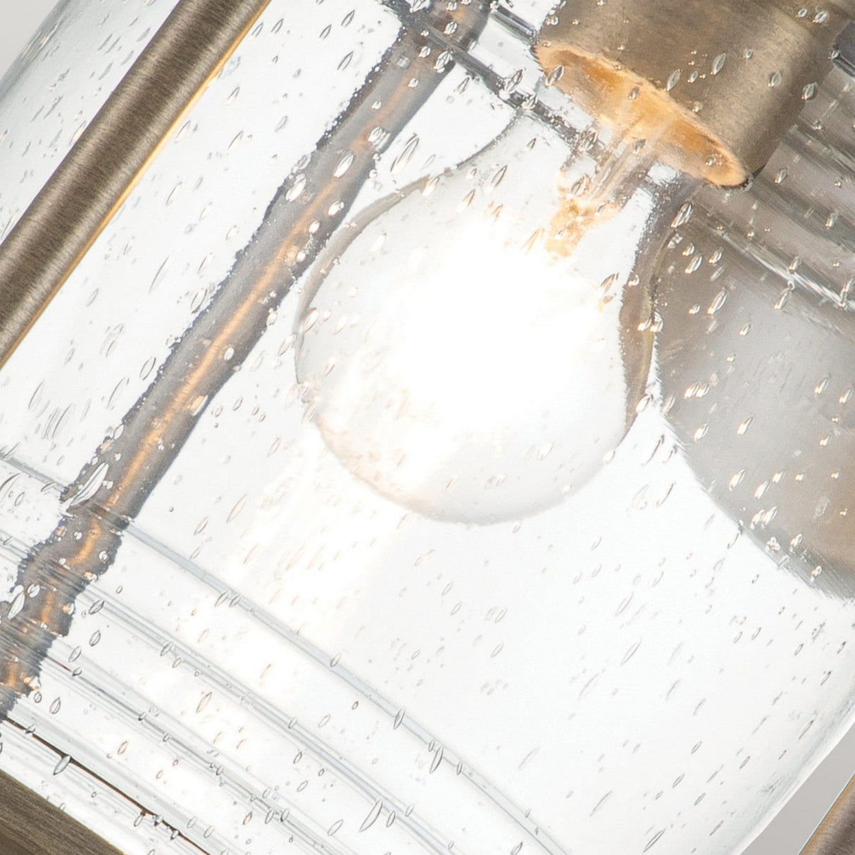 A close-up of an LED Edison-style bulb inside the Ashland Bay Outdoor 1 Light Large Wall Lantern - Burnished Bronze showcases a brass frame. The speckled glass, reminiscent of water droplets, enhances the bulb's warm glow, emphasizing the detailed texture and adding elegance to its burnished bronze finish.
