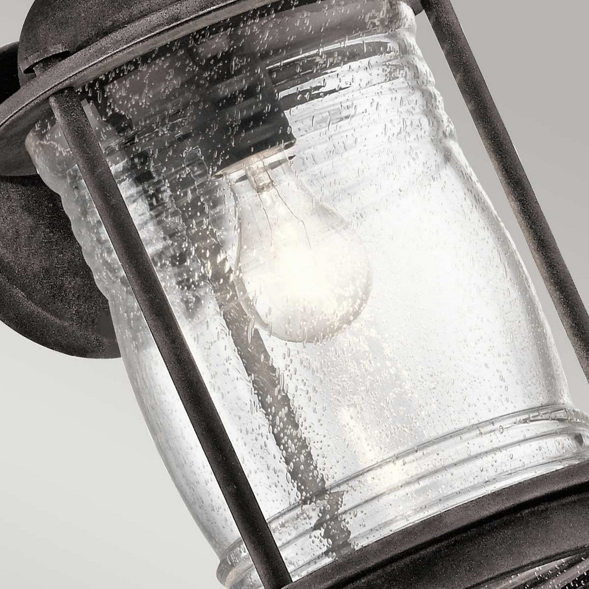 A detailed view of the Ashland Bay Outdoor 1 Light Large Wall Lantern showcases its vintage style, featuring clear, textured glass. Inside, an LED Edison bulb emits a warm glow within a weathered zinc metal frame, all set against a light gray backdrop.